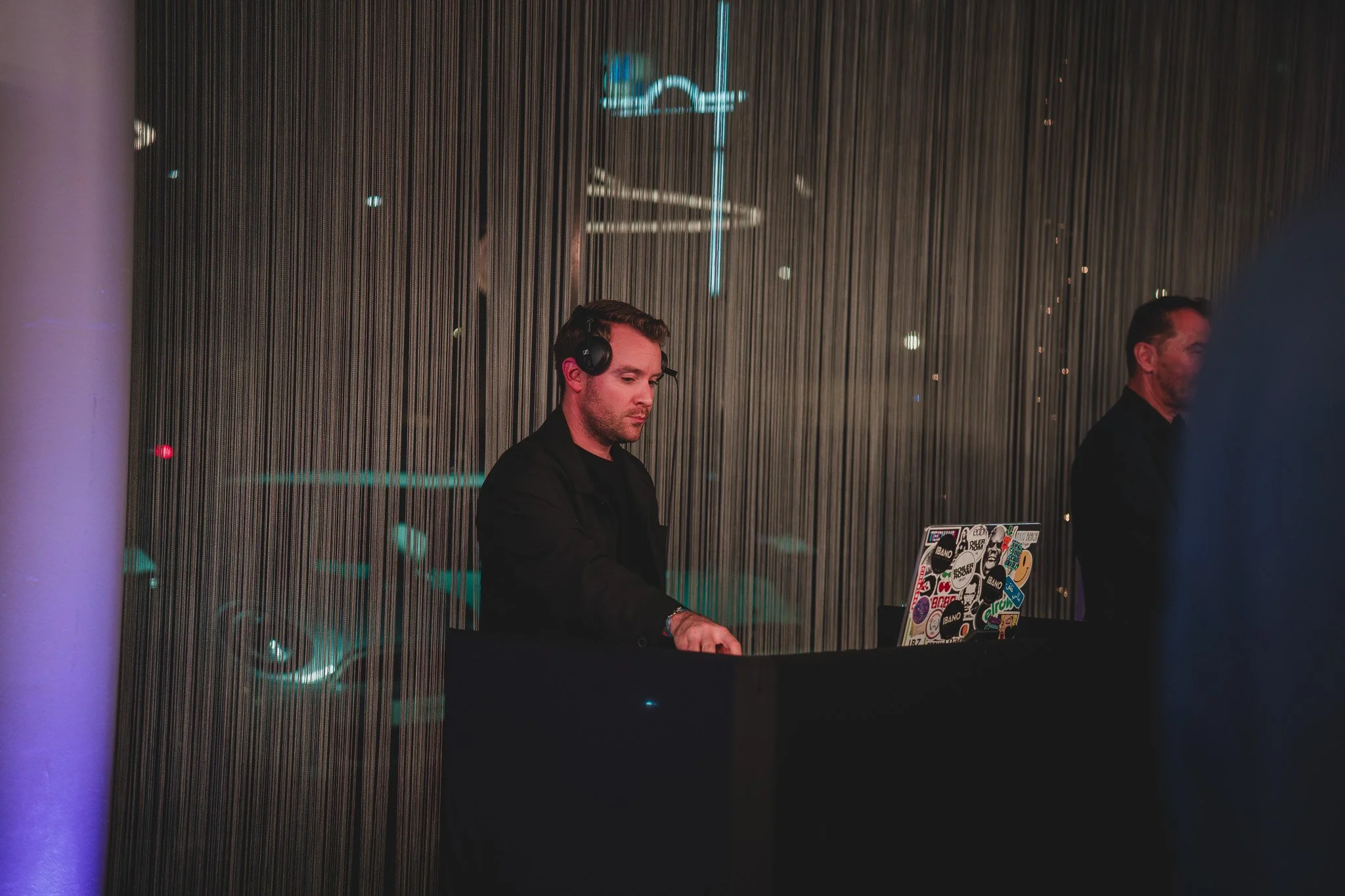 DJ avec écouteurs jouant de la musique derrière un DJ booth dans une ambiance sombre avec un fond de rideau à rayures.