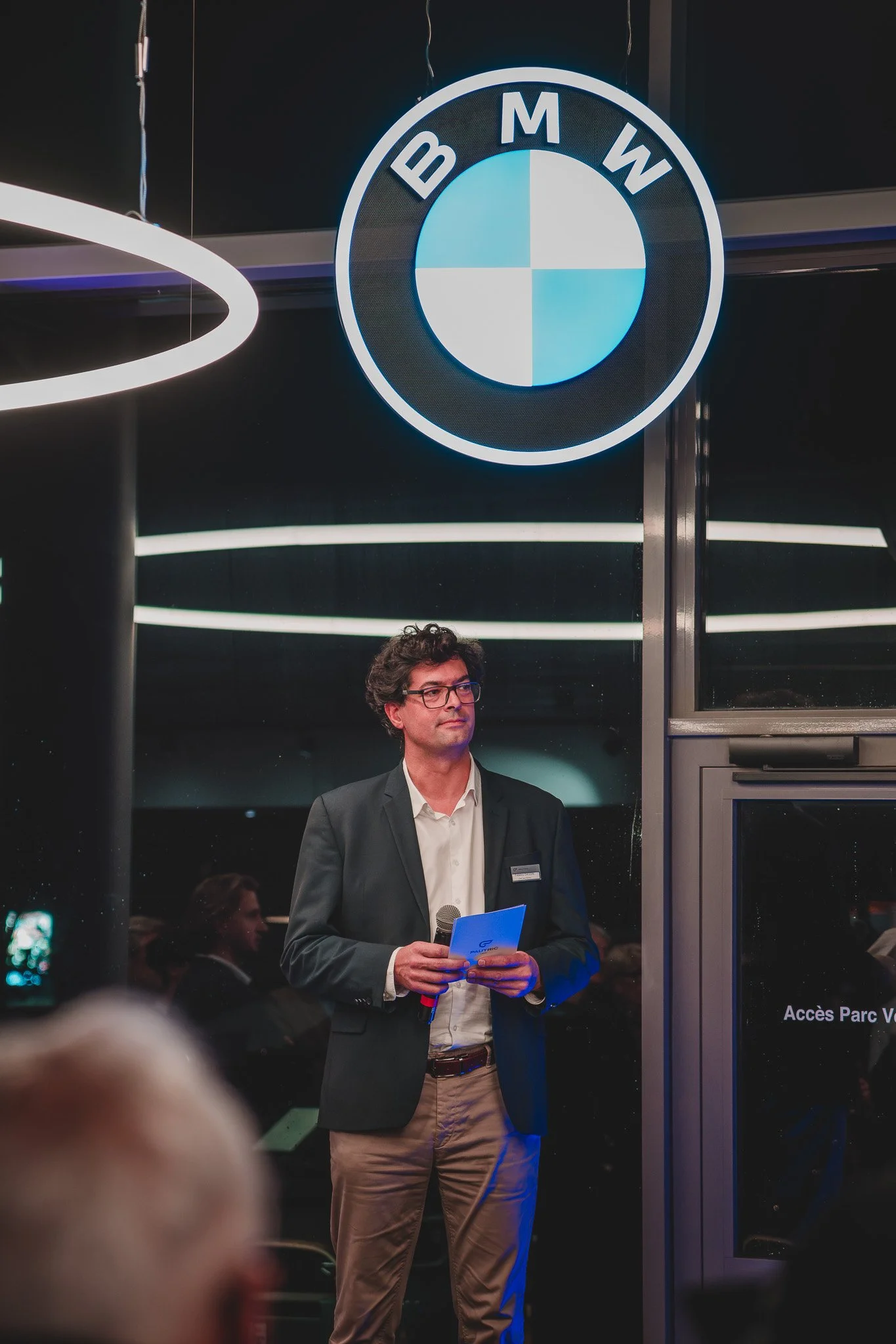 Homme en costume tenu un micro et un document, se tenant sous le logo BMW éclairé, dans un environnement intérieur lors d'un événement ou d'une présentation.