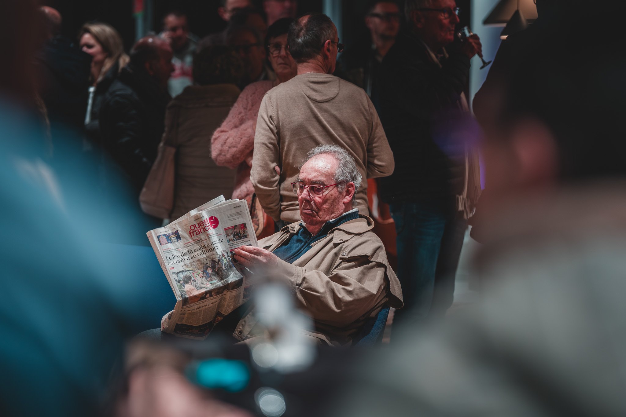 Un homme âgé assis en train de lire un journal dans une pièce bondée de gens qui discutent.