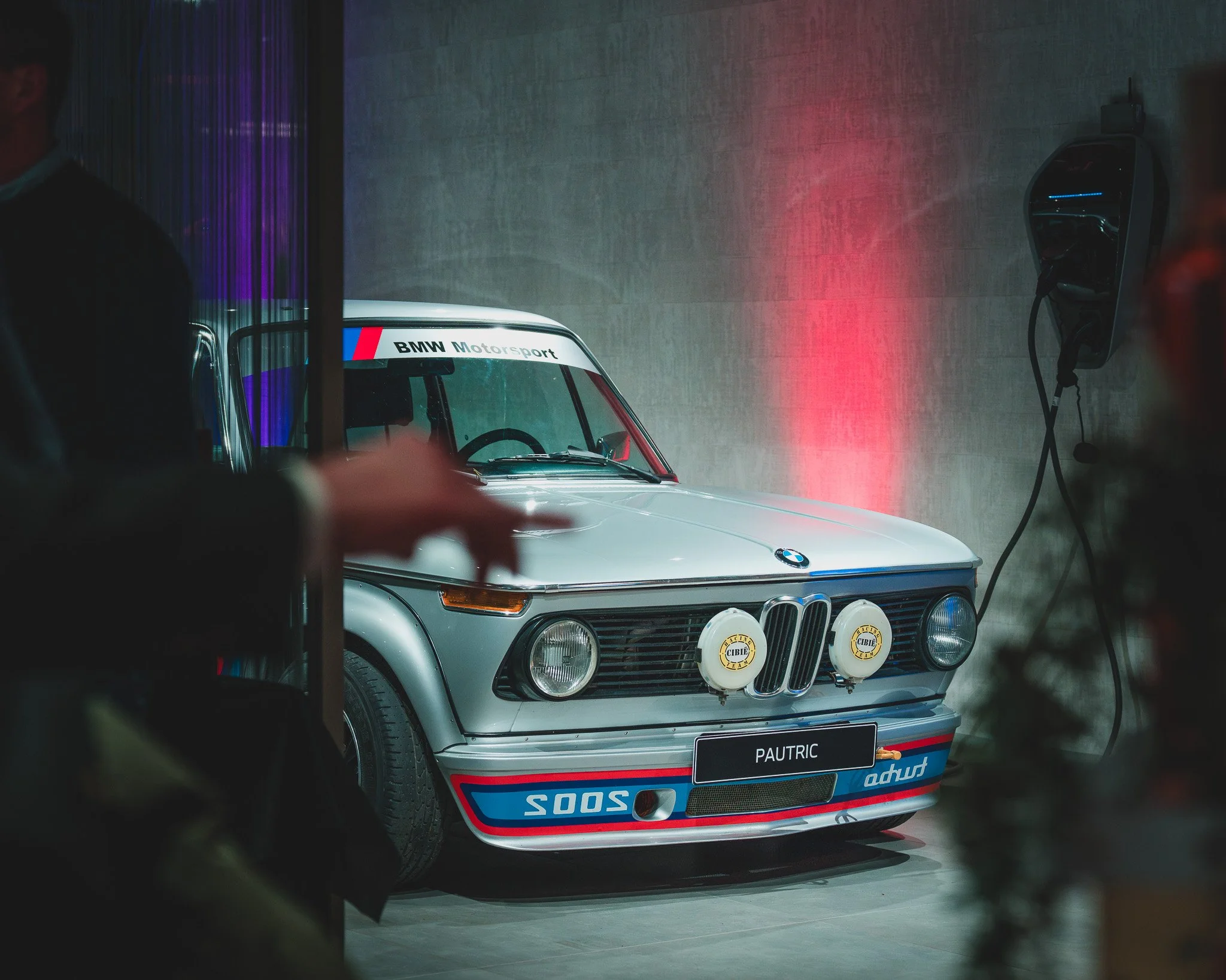 Une vieille voiture de course BMW, de couleur blanche avec des accents bleus et rouges, exposée dans une salle avec un éclairage tamisé et des néons violets et rouges.