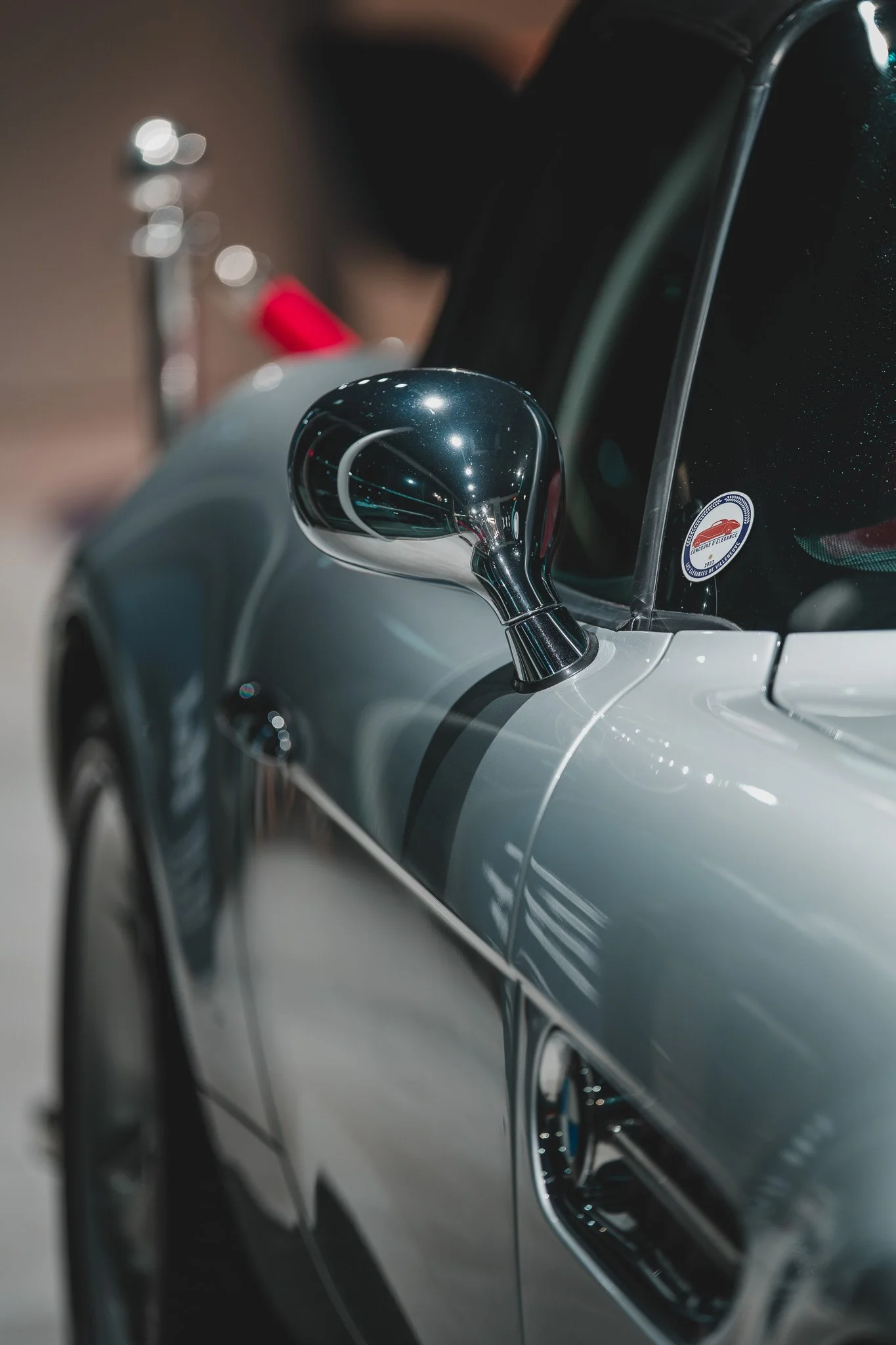 Photo en gros plan d'un rétroviseur d'une voiture de sport de couleur gris argenté, avec un badge ou une plaque près du pare-brise. L'image est bien éclairée avec un reflet sur la surface du rétroviseur.