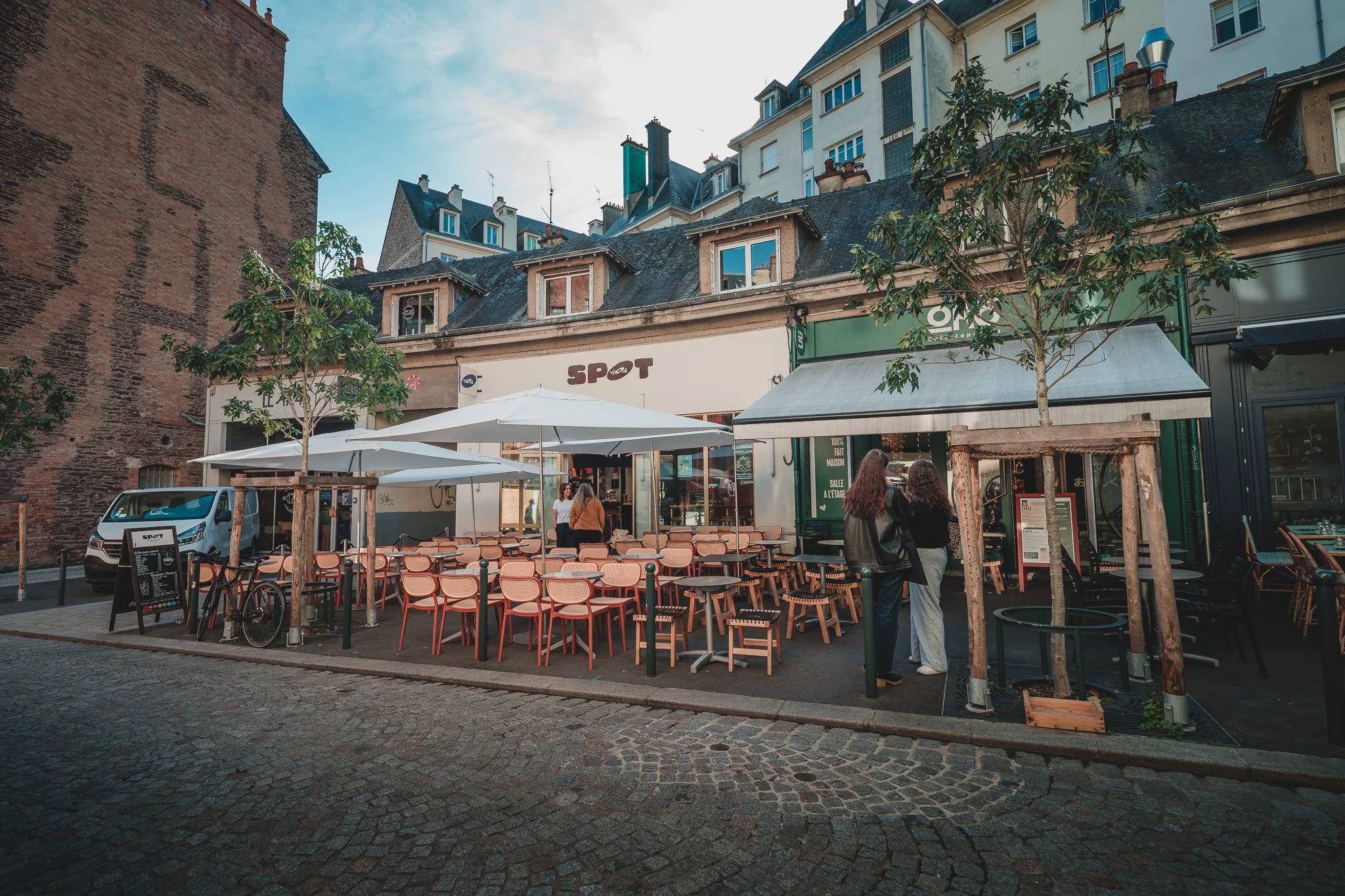 Terrasse de restaurant avec tables, chaises et parasols, deux femmes discutent devant un établissement nommé 'SPOT' dans une rue pavée urbaine, avec bâtiments en arrière-plan.