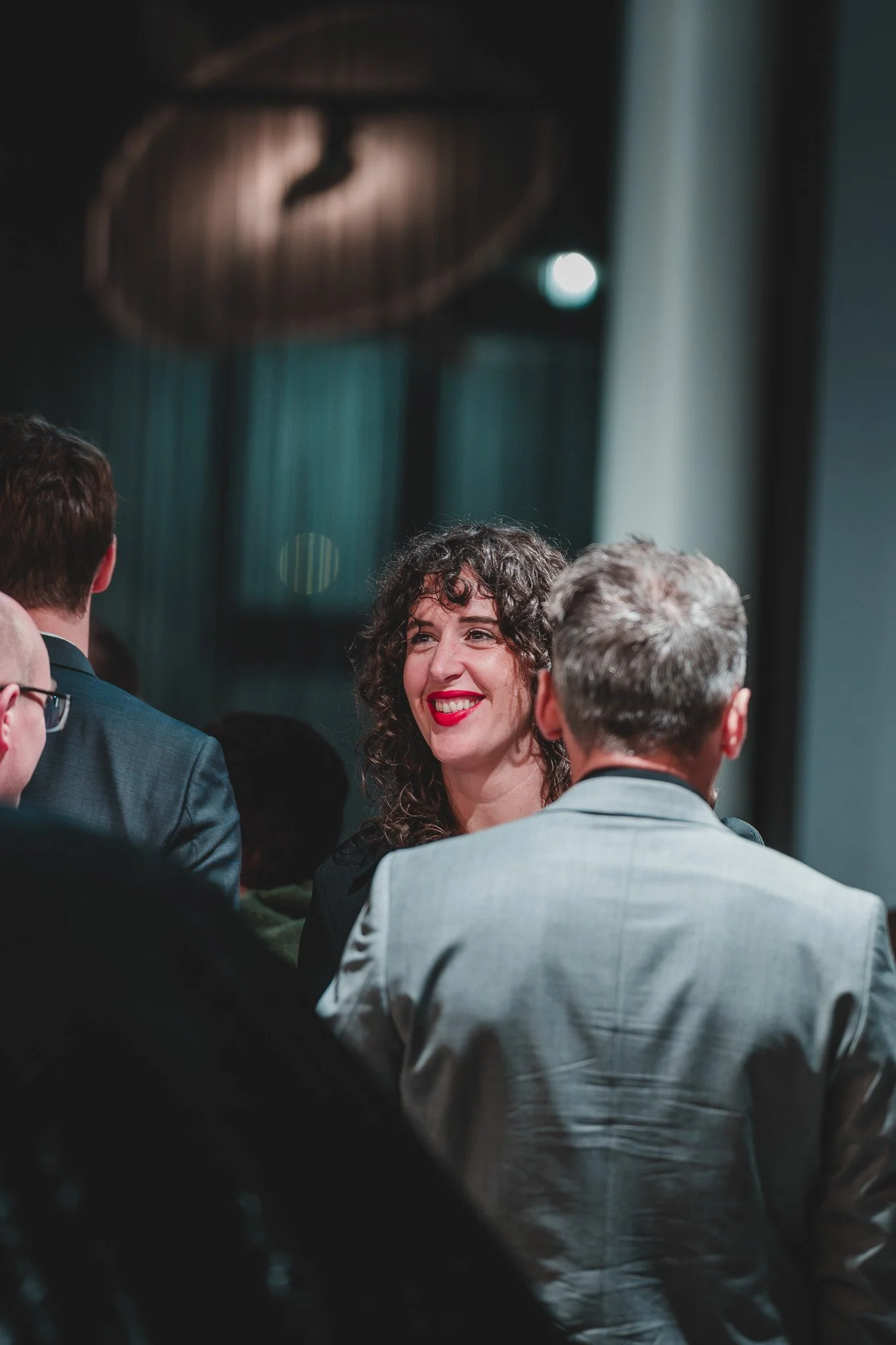 Une femme souriante avec des cheveux bouclés, portant un rouge à lèvres rouge, parlait dans une cérémonie ou un événement, entourée de plusieurs hommes en costume.