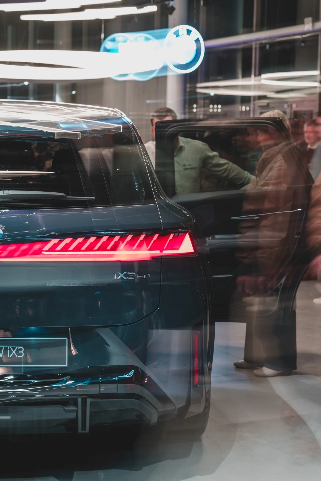 Une voiture électrique noire de marque BMW, modèle iX3, exposée dans un salon auto, avec des personnes qui la regardent et discutent autour.