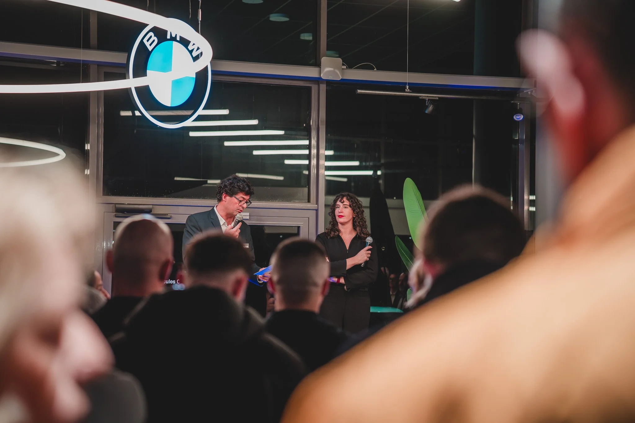 Deux personnes parlent lors d'une présentation ou d'une conférence dans une salle moderne avec un logo BMW en arrière-plan. Un homme en costume tient un micros et regarde ses notes, une femme en noir tient un micro et regarde vers l'auditoire.