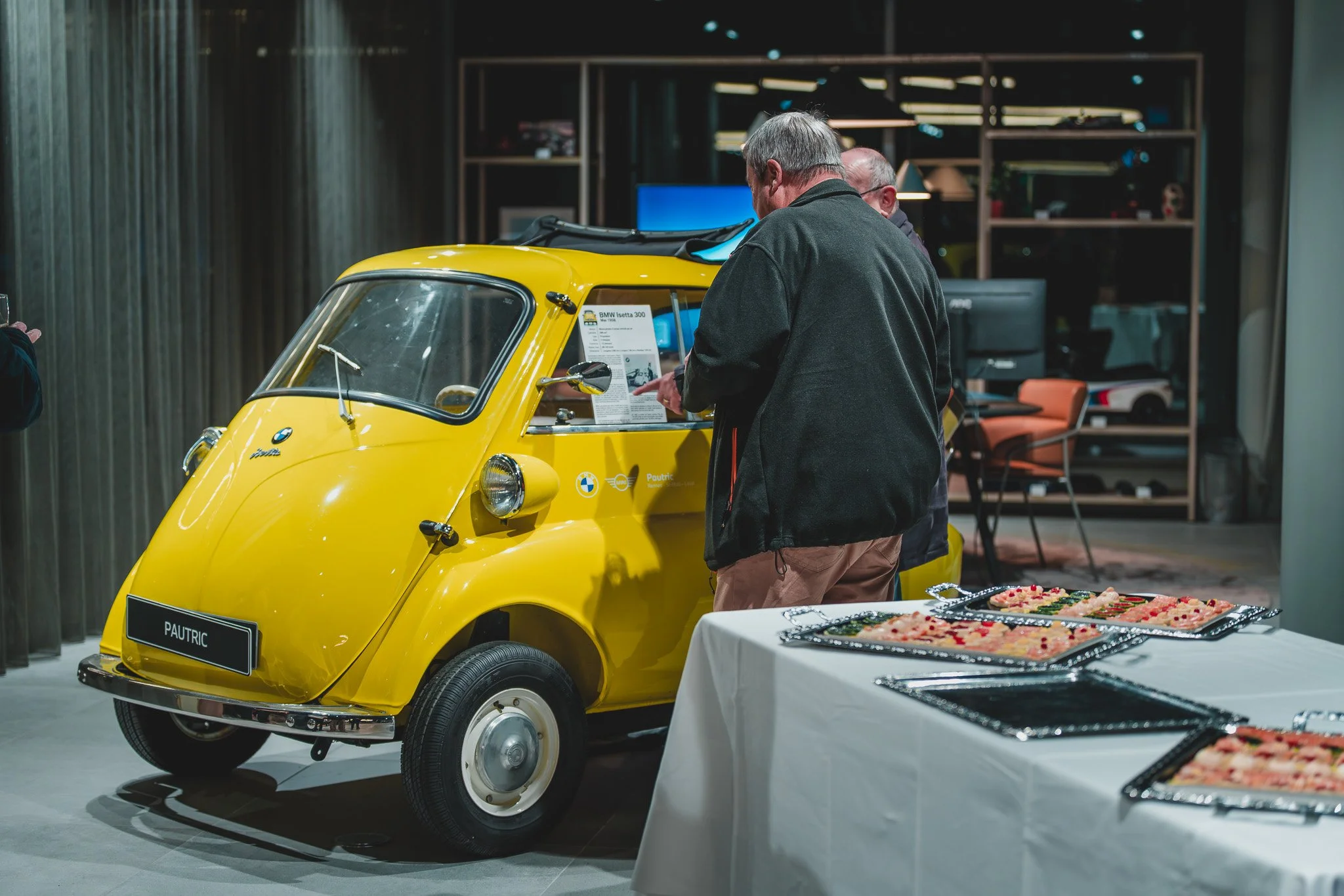 Deux hommes regardent un véhicule jaune, une voiture à trois roues, dans une exposition ou un musée, avec des tables de nourriture à côté.
