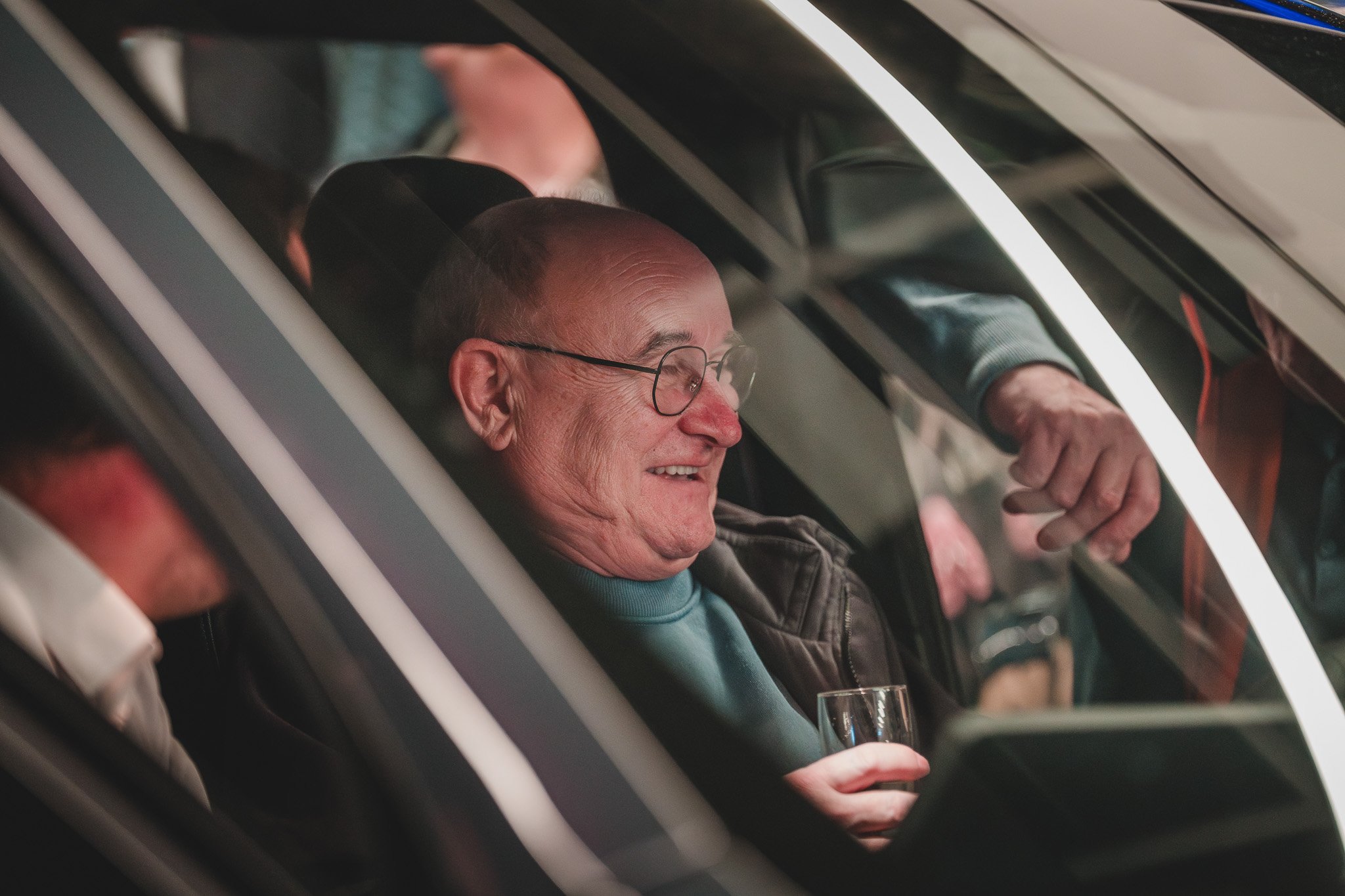 Un homme âgé portant des lunettes sourit en tenant un verre, assis dans une cabine de simulation de vol ou un cinéma en réalité virtuelle, entouré d'autres personnes.