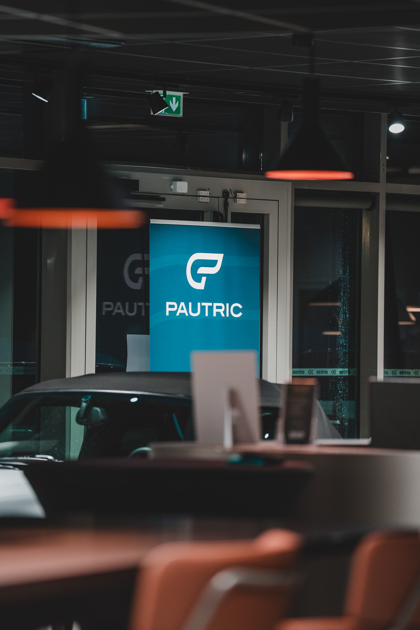 Une publicité pour la marque PAUTRIC à l'intérieur d'une salle ou d'un hall, avec des meubles et une voiture visible en arrière-plan, dans un environnement intérieur moderne.