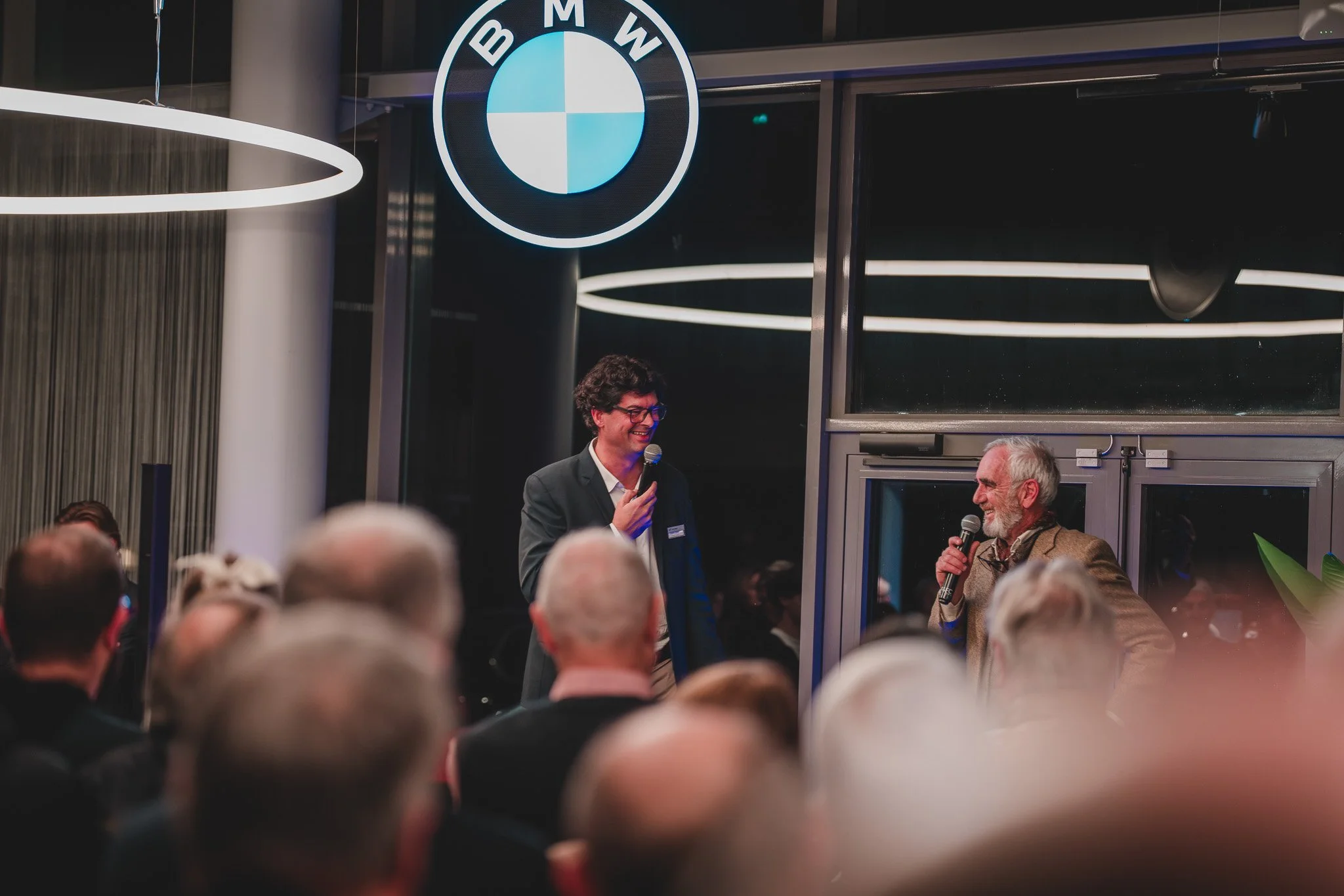 Deux hommes parlent lors d'une conférence ou d'un événement, debout devant un public. Il y a un logo BMW en arrière-plan, dans une salle moderne avec un éclairage contemporain.