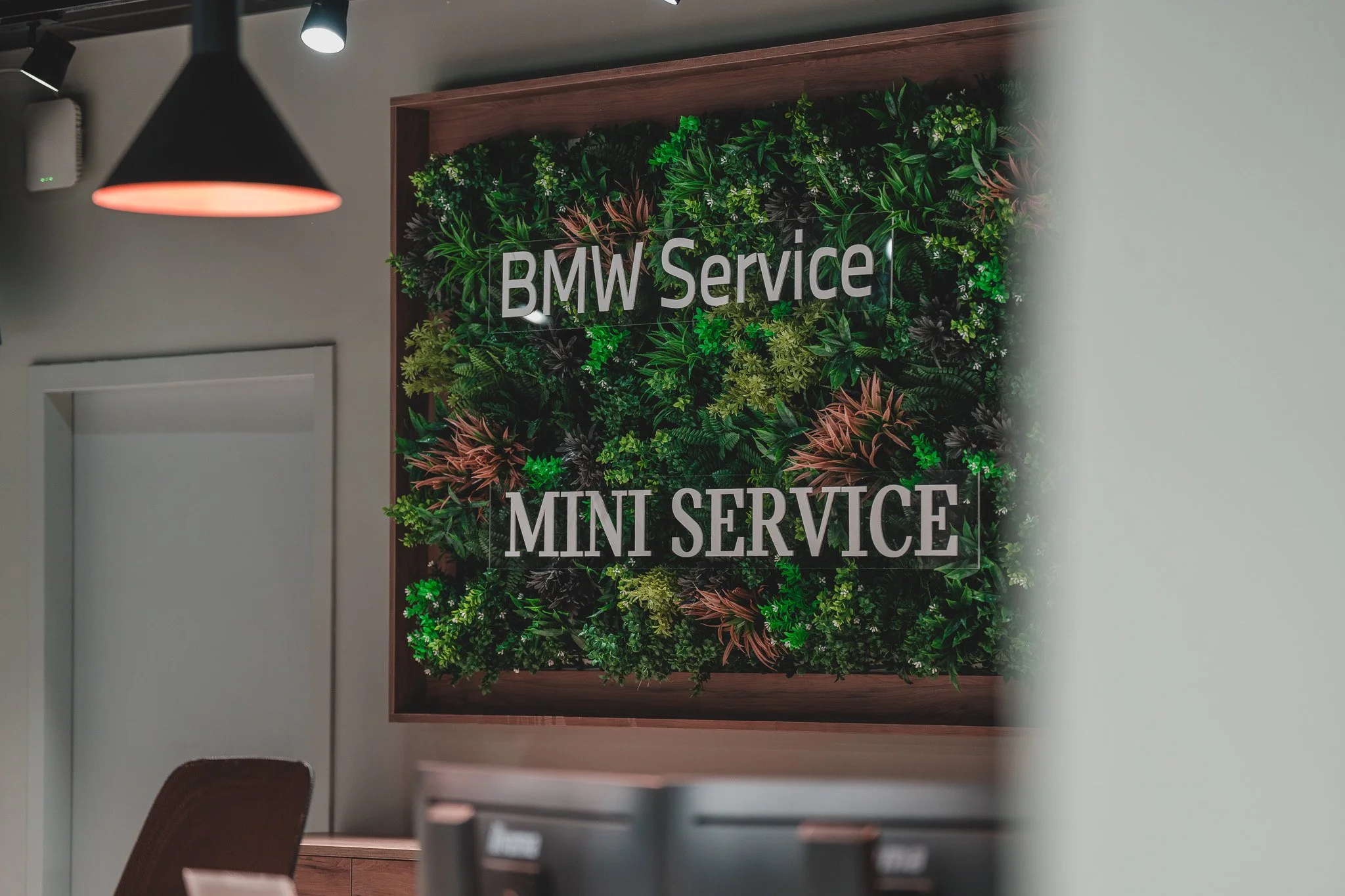 Panneau vert avec écritures blanches indiquant 'BMW Service' et 'MINI SERVICE', entouré de végétation verte. Vu à l'intérieur d'un espace de service automobile.