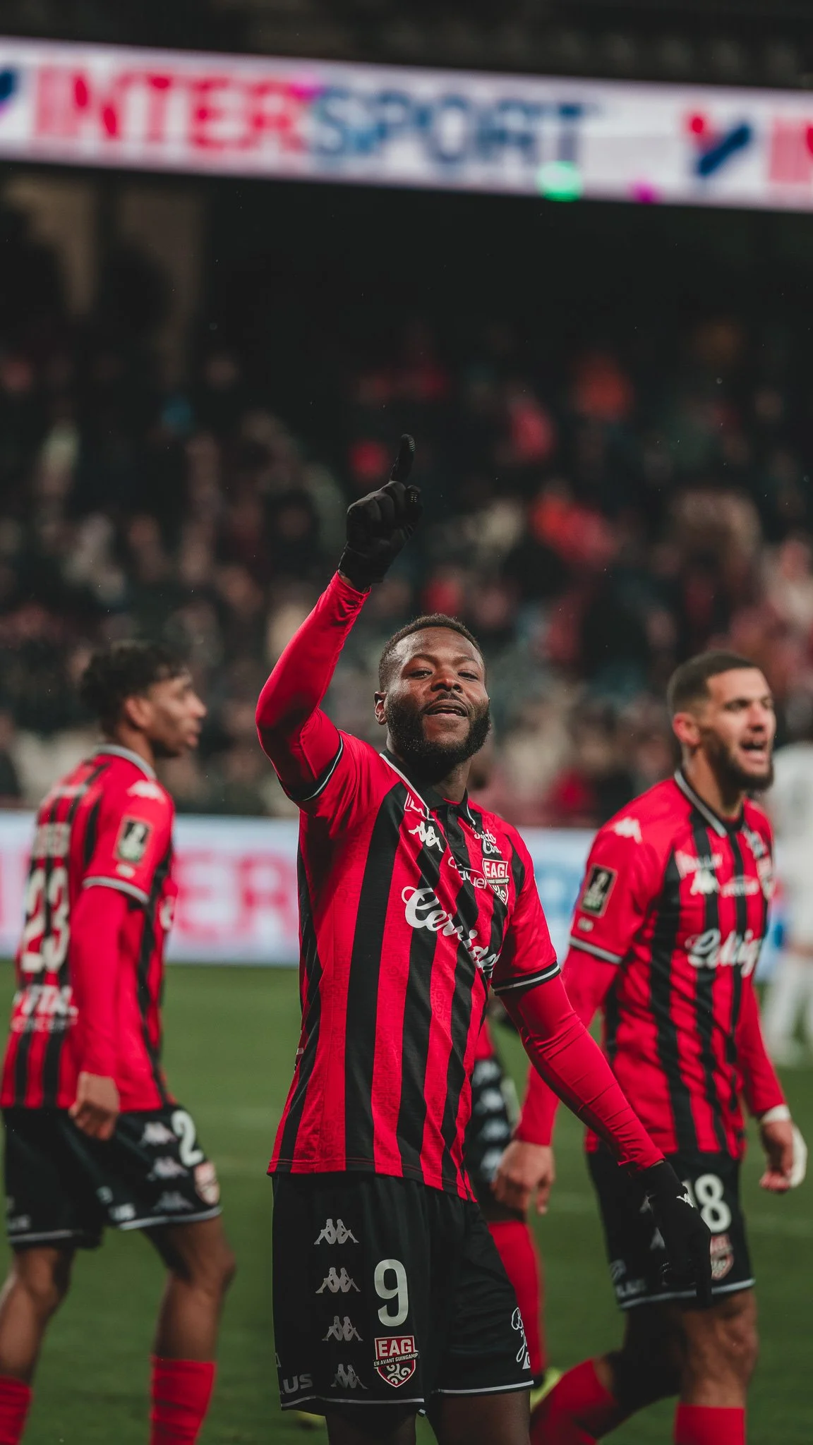 Des joueurs de football en train de célébrer un but sur le terrain, portant des maillots rouges et noirs avec le logo de leur équipe, dans un stade plein de spectateurs.