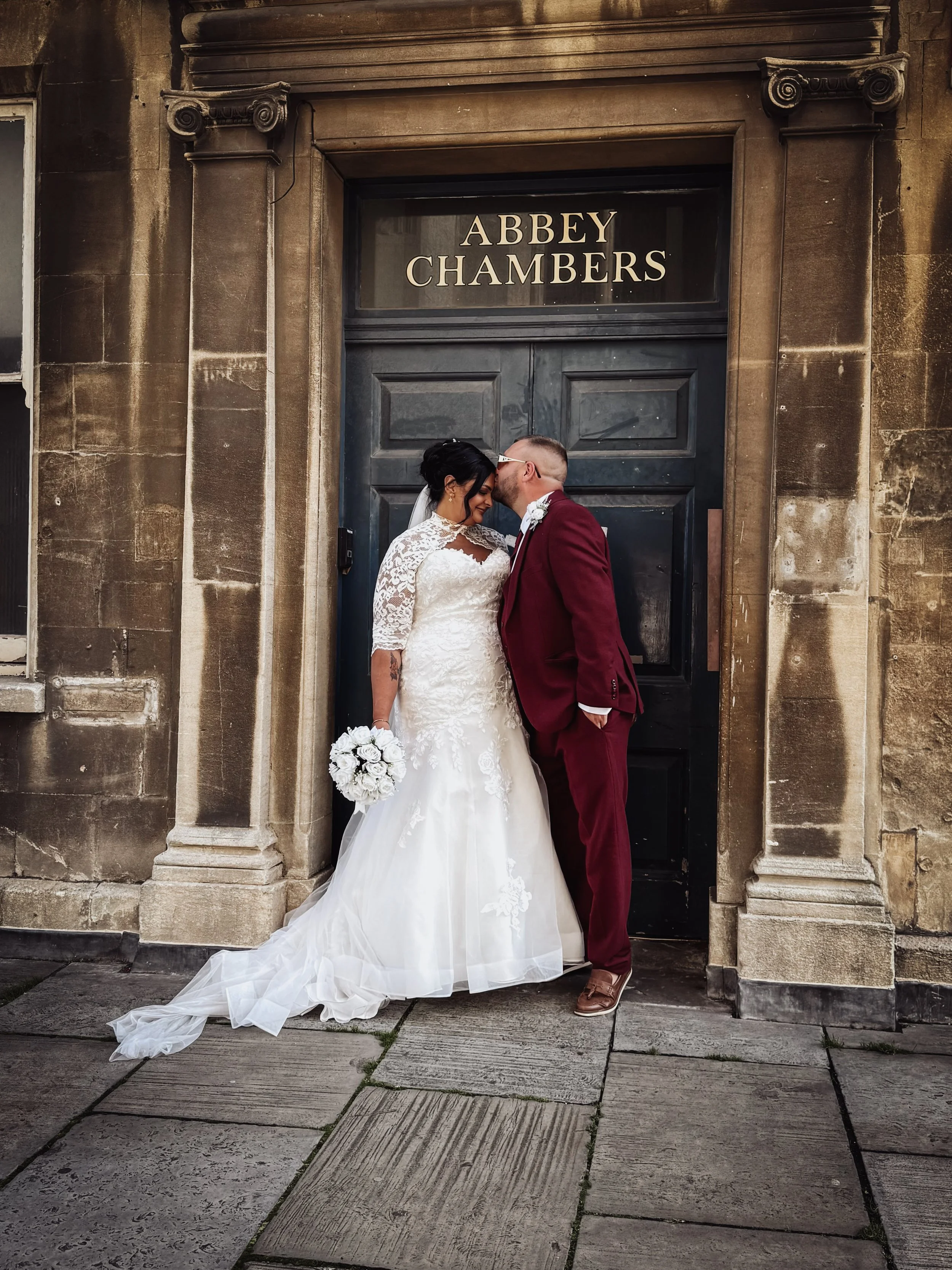 A newlywed couple shares a kiss outside the Abbey Chambers, with stone columns surrounding a black door.