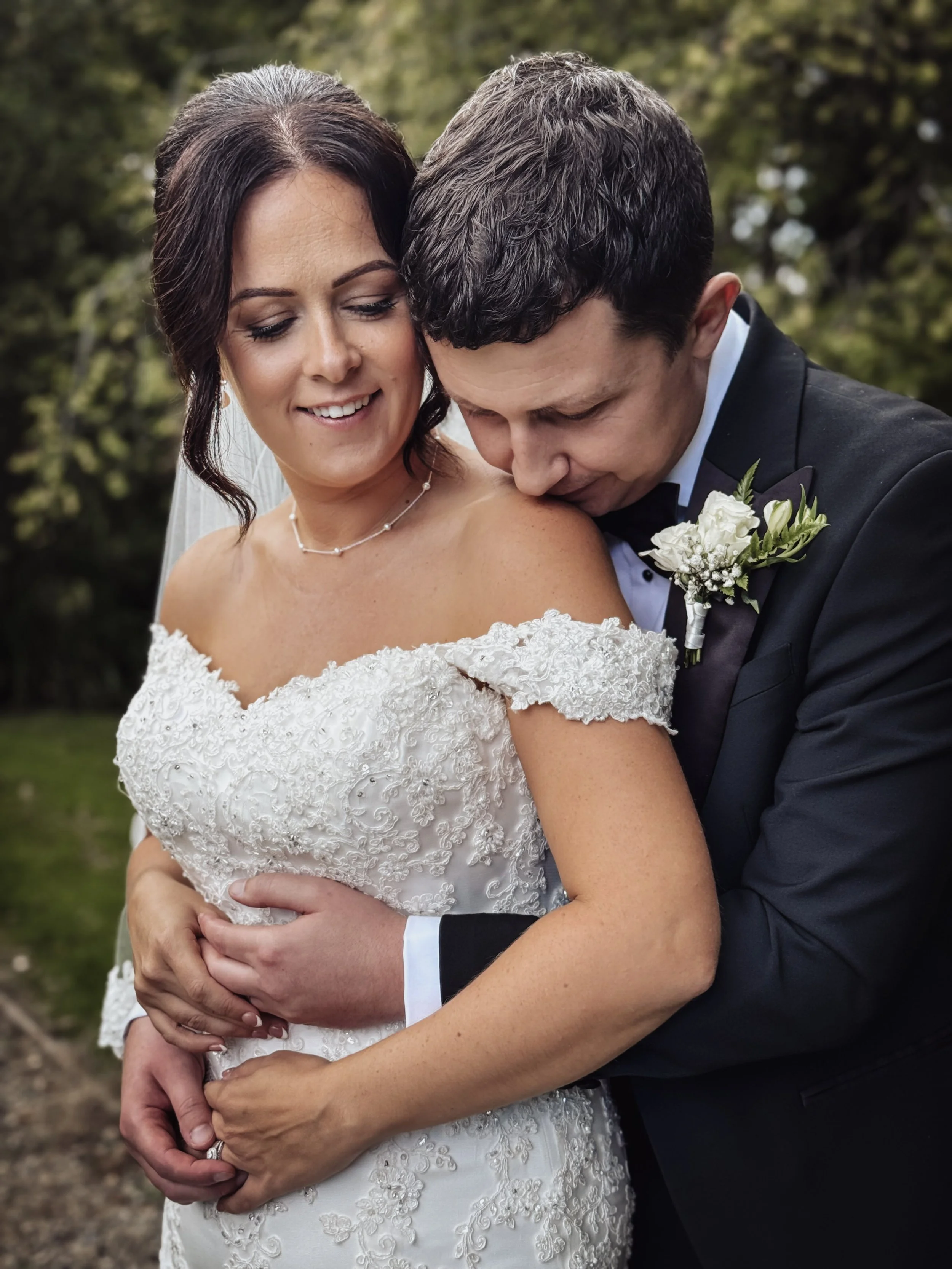 A bride in a white lace wedding gown and a groom in a black tuxedo embrace outdoors, smiling with trees in the background.