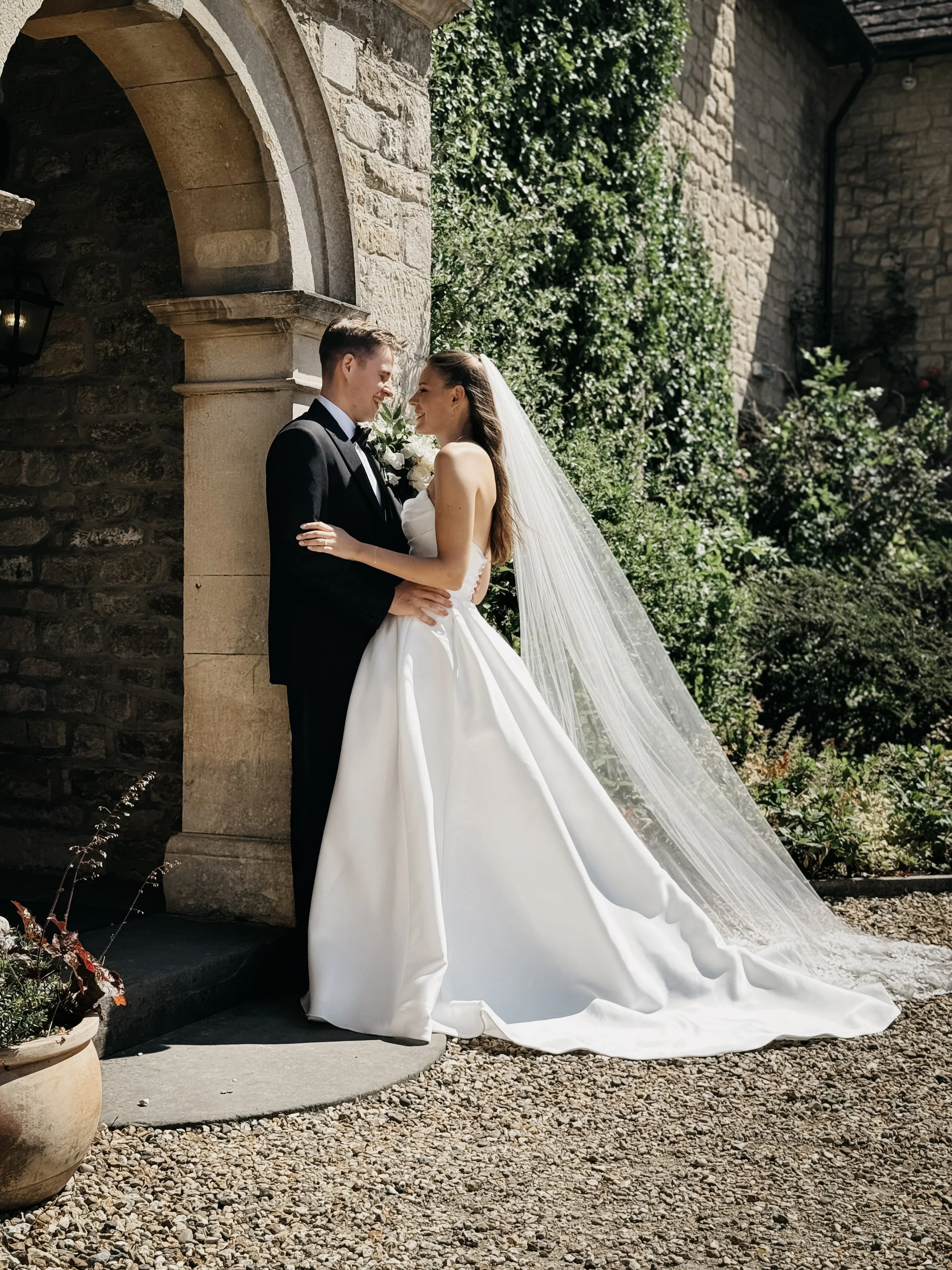 A bride and groom in wedding attire sharing a kiss outdoors, with the bride in a white gown and veil, and the groom in a black tuxedo, near a stone building with greenery.