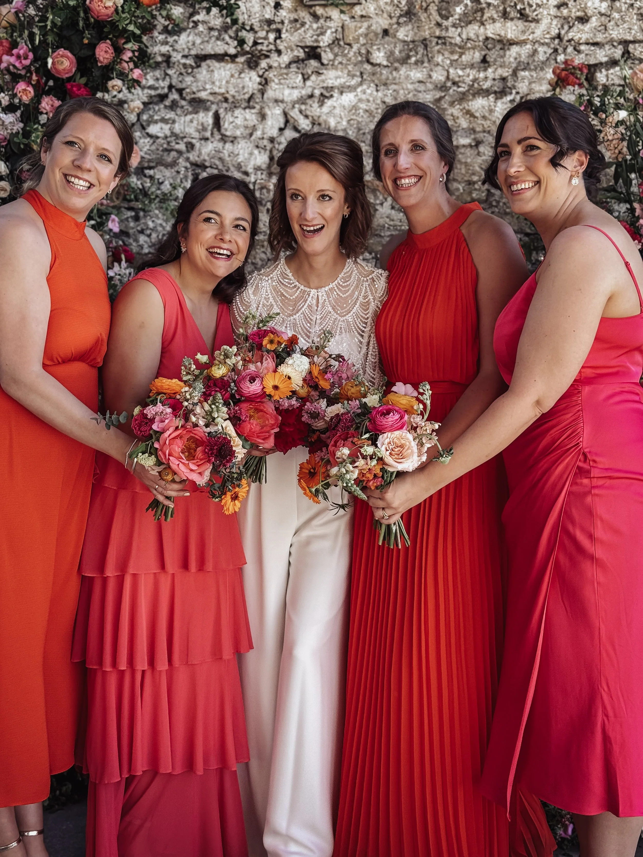 Bride in a white dress holding a bouquet of pink, orange, and red flowers, surrounded by five women in bright pink and orange dresses holding bouquets, standing against a stone wall with floral decorations.