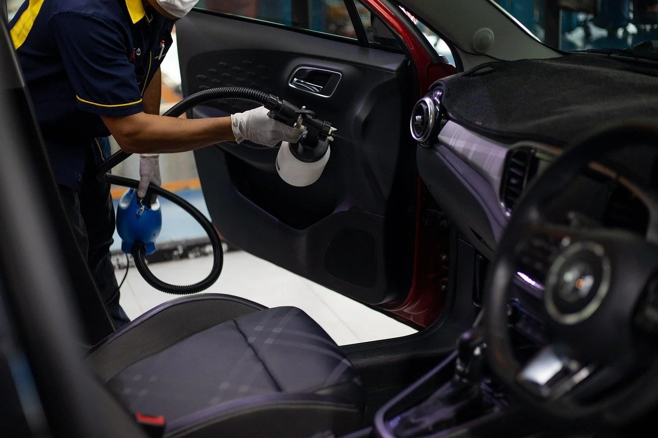 A person cleaning the interior of a red car using a spray bottle and a handheld device with a hose.