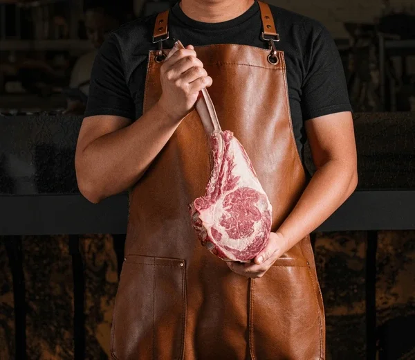 Person wearing a leather apron holding a large cut of raw meat.