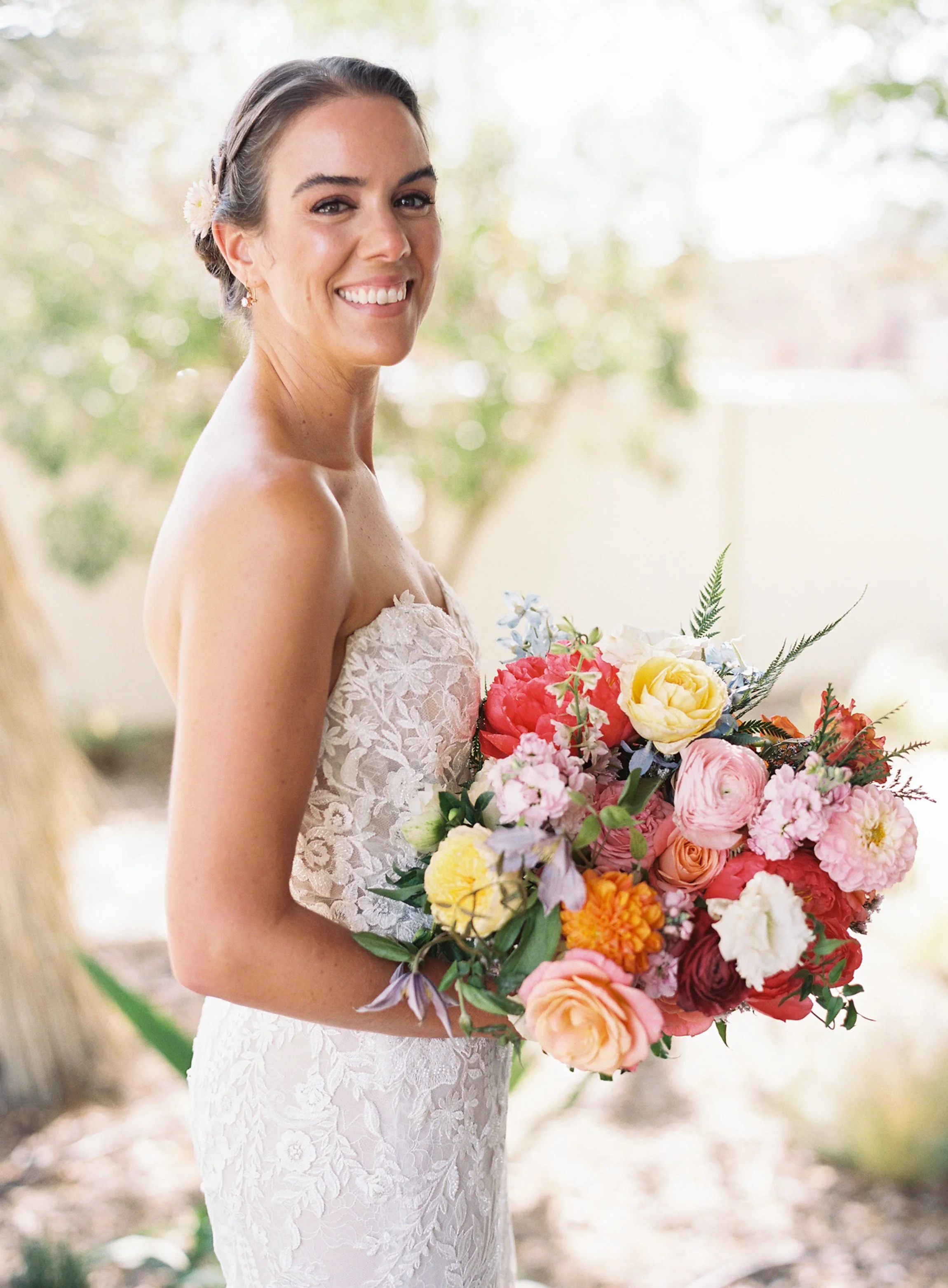 A bride with dark hair styled in a braid, wearing a strapless lace wedding dress, holding a colorful bouquet of flowers, standing outdoors with greenery in the background, smiling at the camera.