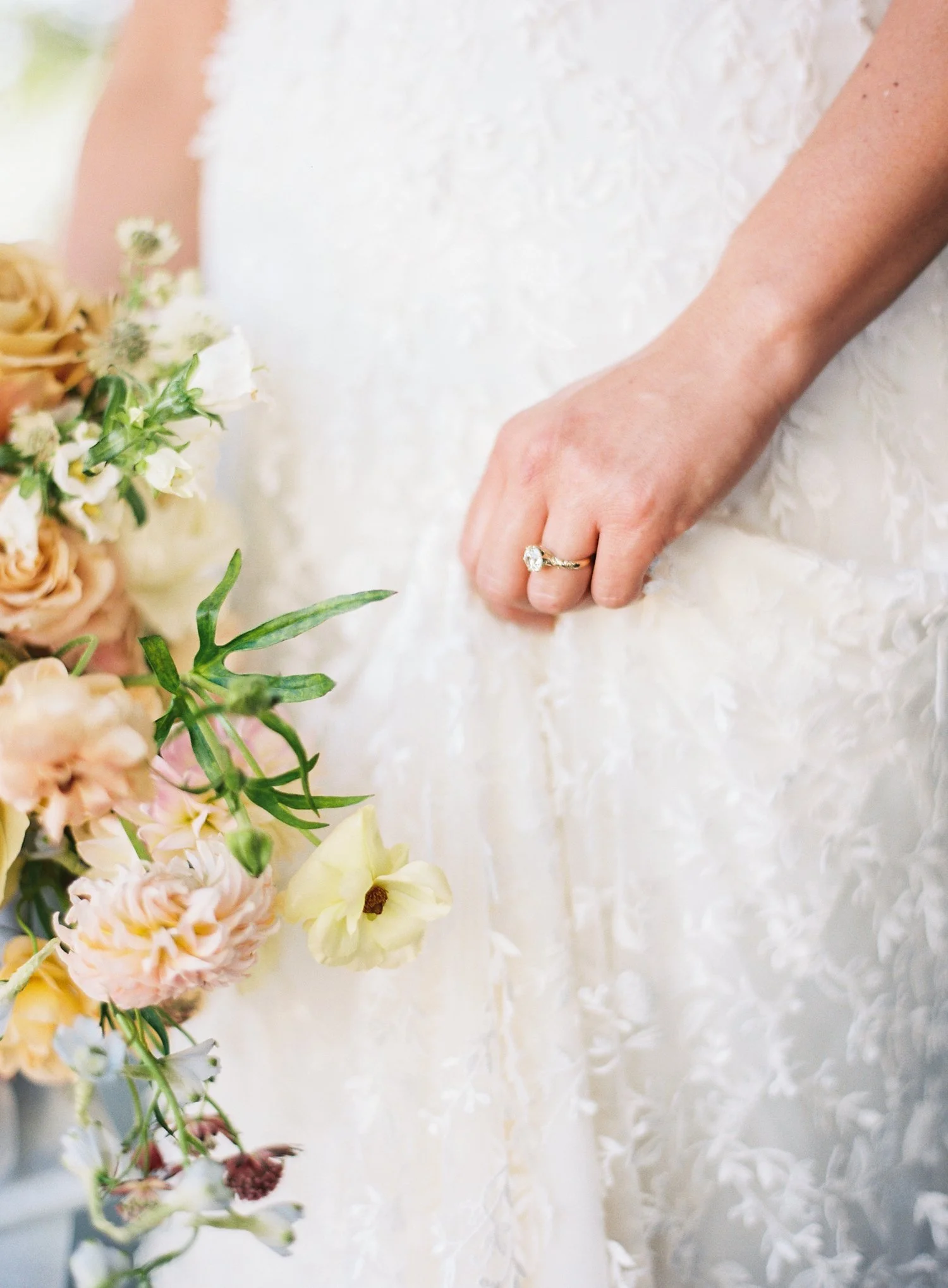 Close-up of a bride holding her wedding dress, wearing a diamond ring, with a bouquet of pastel-colored flowers.