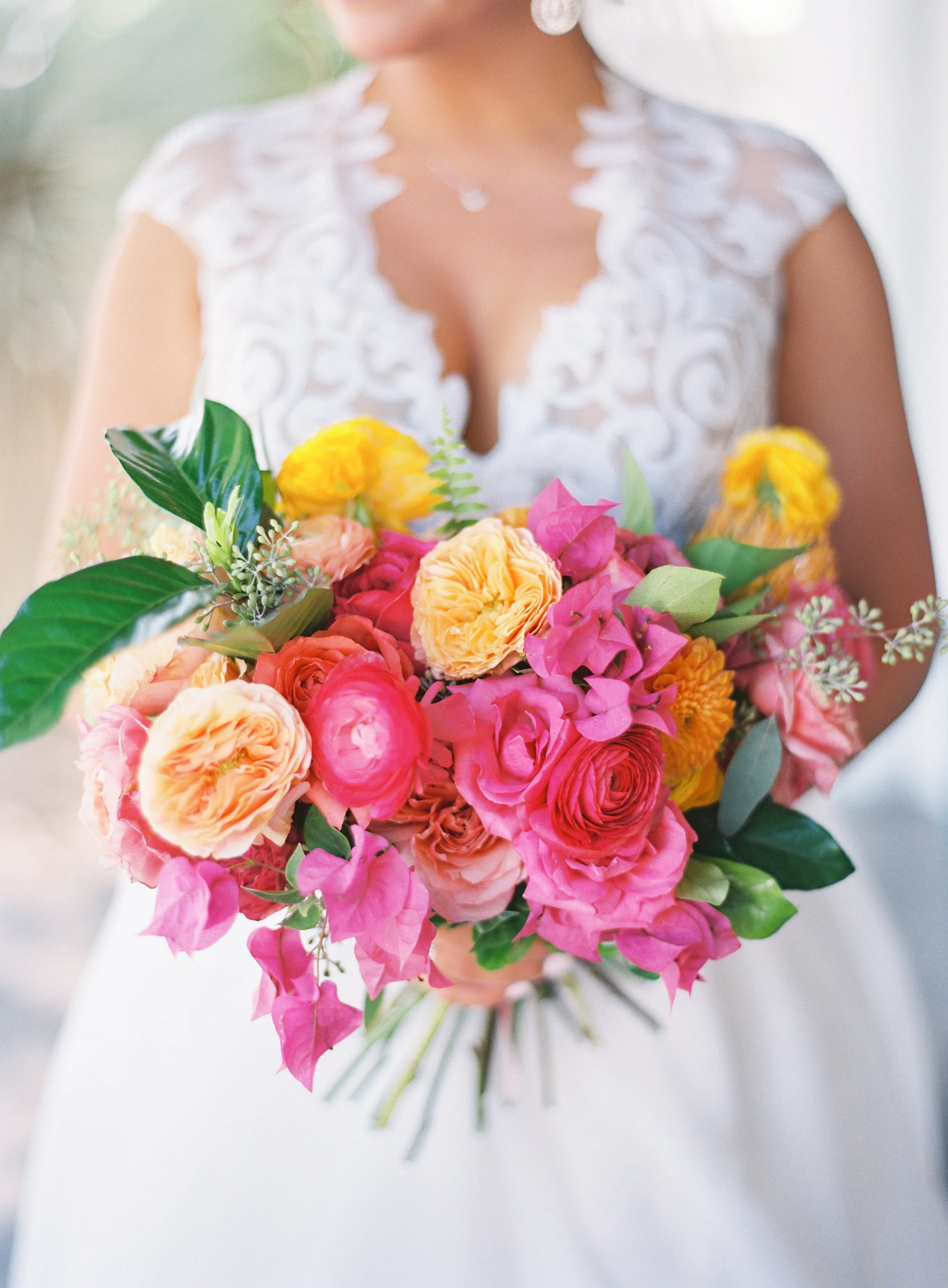 Bride holding a colorful bouquet of pink, yellow, peach, and orange flowers with green leaves.