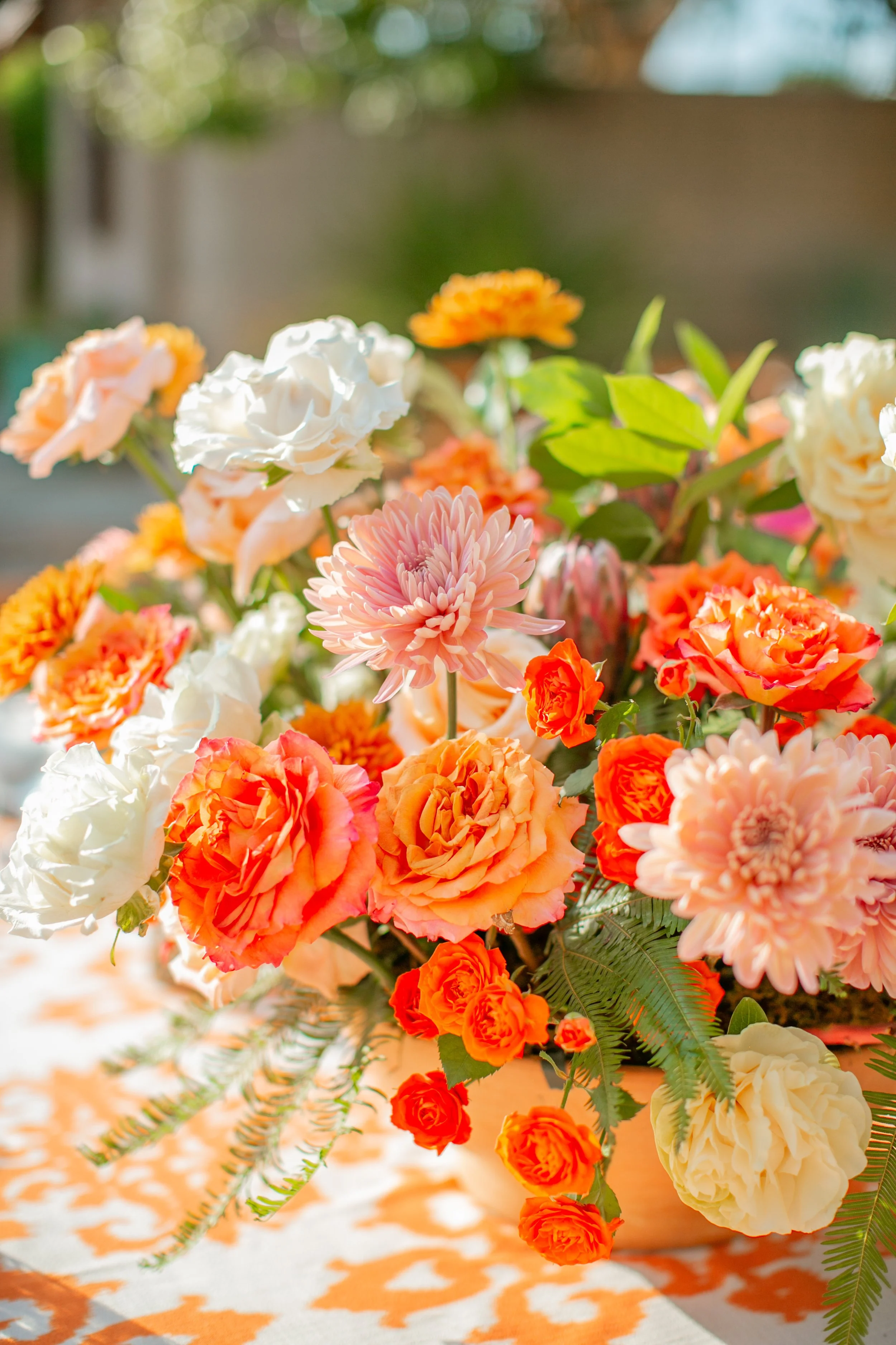 A colorful bouquet of various flowers, including pink, orange, and white blooms, with green foliage, placed on a patterned orange and white tablecloth outdoors.