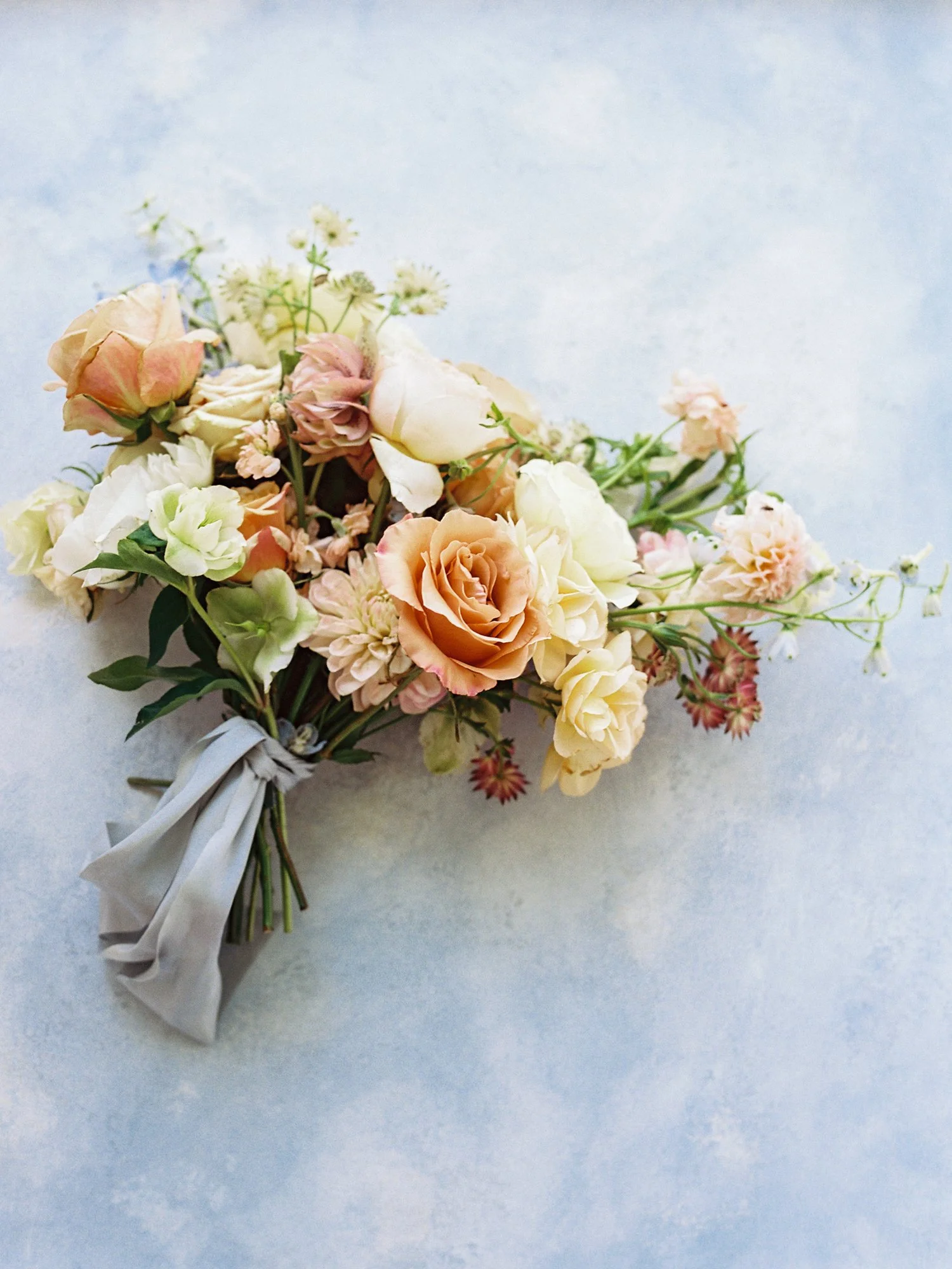 A bouquet of pastel-colored roses and other flowers tied with a gray ribbon on a light textured background.