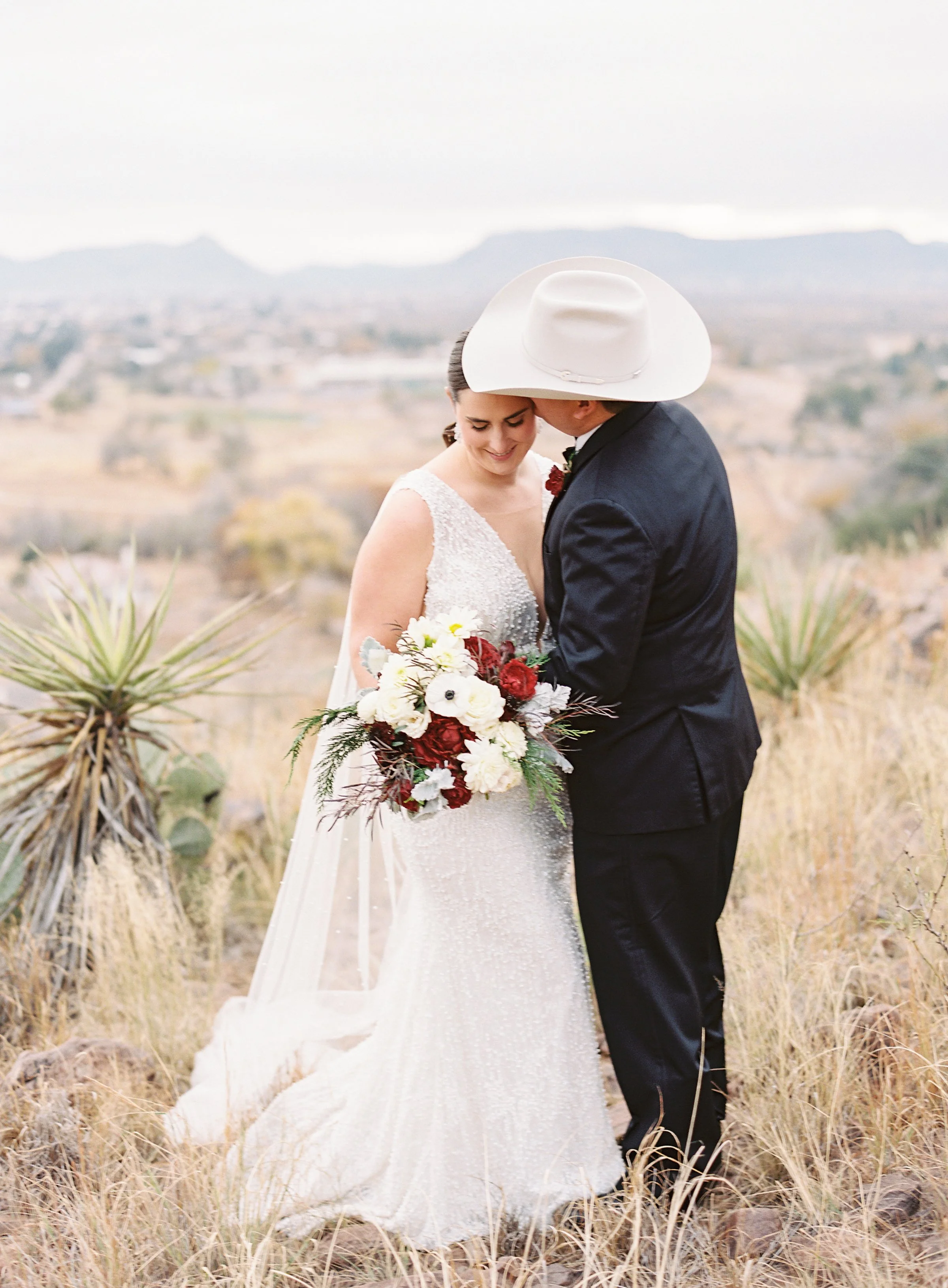 A bride and groom sharing a kiss outdoors in a desert landscape, with the bride holding a bouquet of red and white flowers.