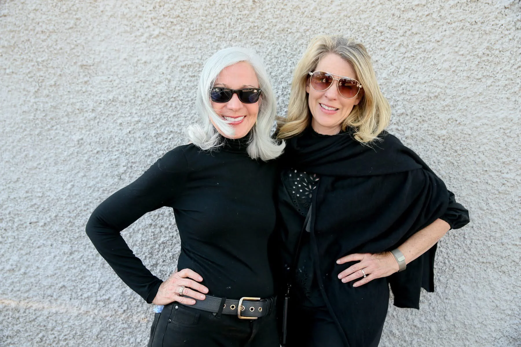 Two women with blonde hair wearing black clothing and sunglasses standing against a light-colored textured wall, smiling with arms around each other.