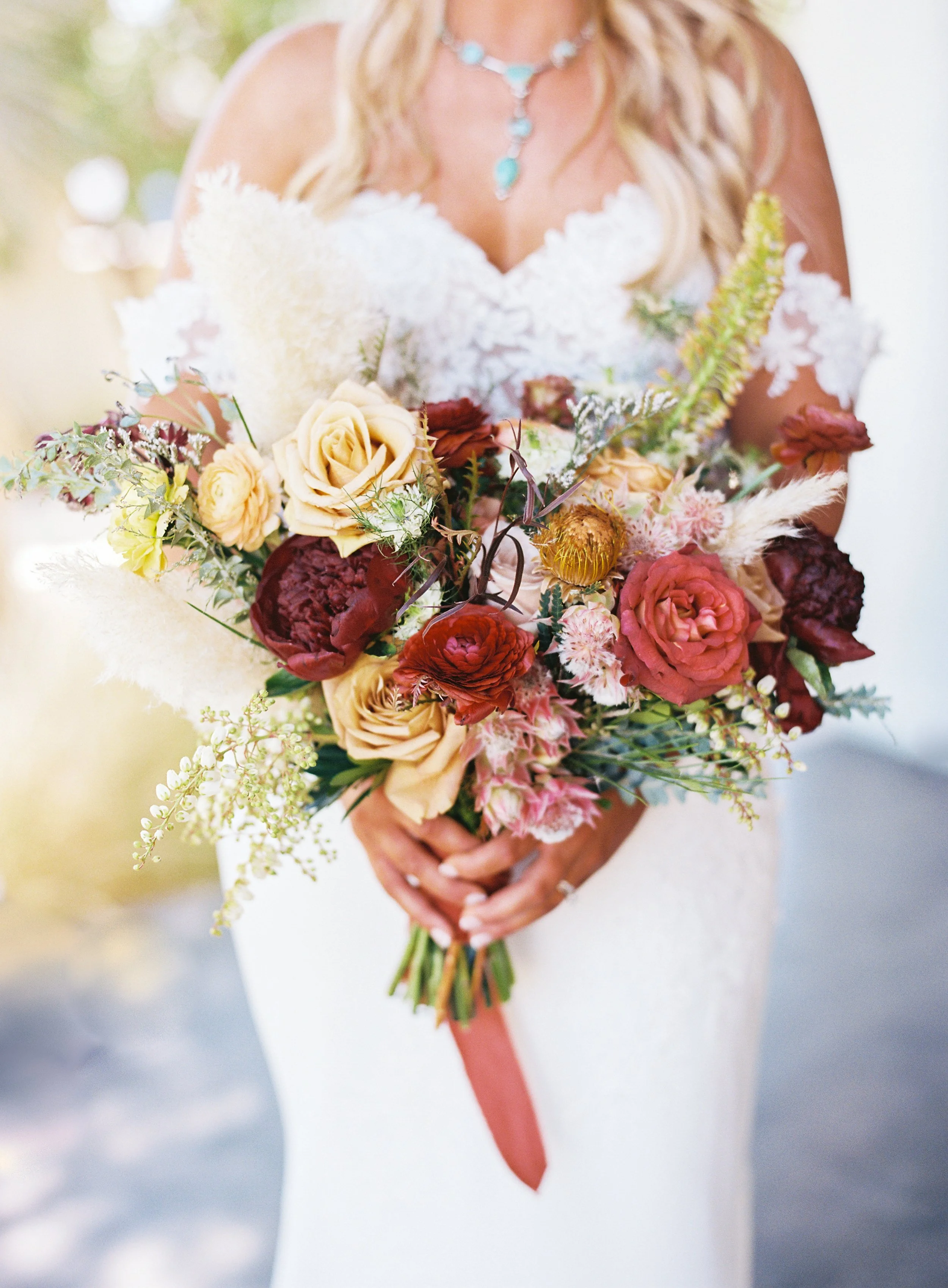 A bride holding a large bouquet of flowers, including roses, peonies, and various greenery, in front of her white wedding dress.