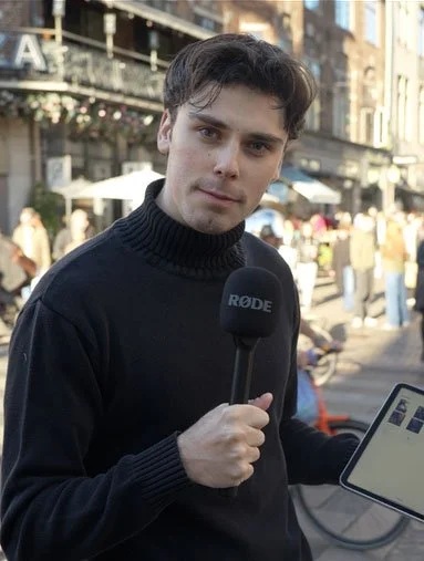 Young man with dark hair, wearing a black turtleneck sweater, holding a microphone with the RØDE logo, standing outdoors in a city setting with buildings, people, and outdoor seating in the background.