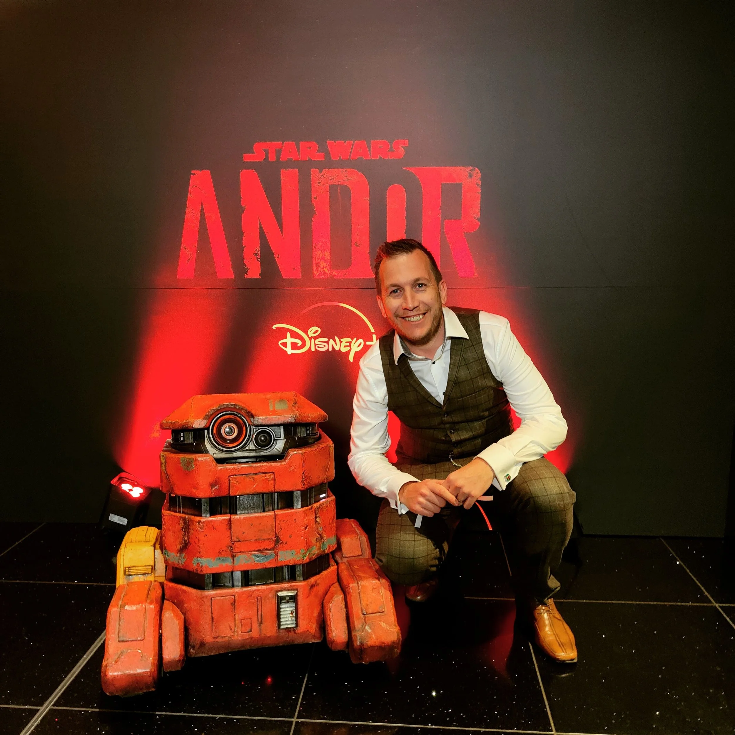 A man in formal attire kneeling next to a large red robot prop from Disney's Star Wars 'Andor' series, in front of a black wall with the 'Andor' and Disney logos illuminated in red.