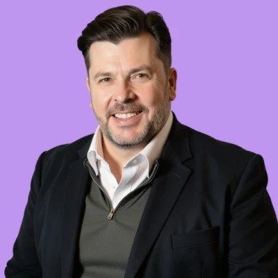 Karl Wood - Director and founder of WINCHR. A man with dark hair and a beard smiling, wearing a black blazer over a gray sweater with a white collared shirt, against a purple background. Hospitality HR services near me.