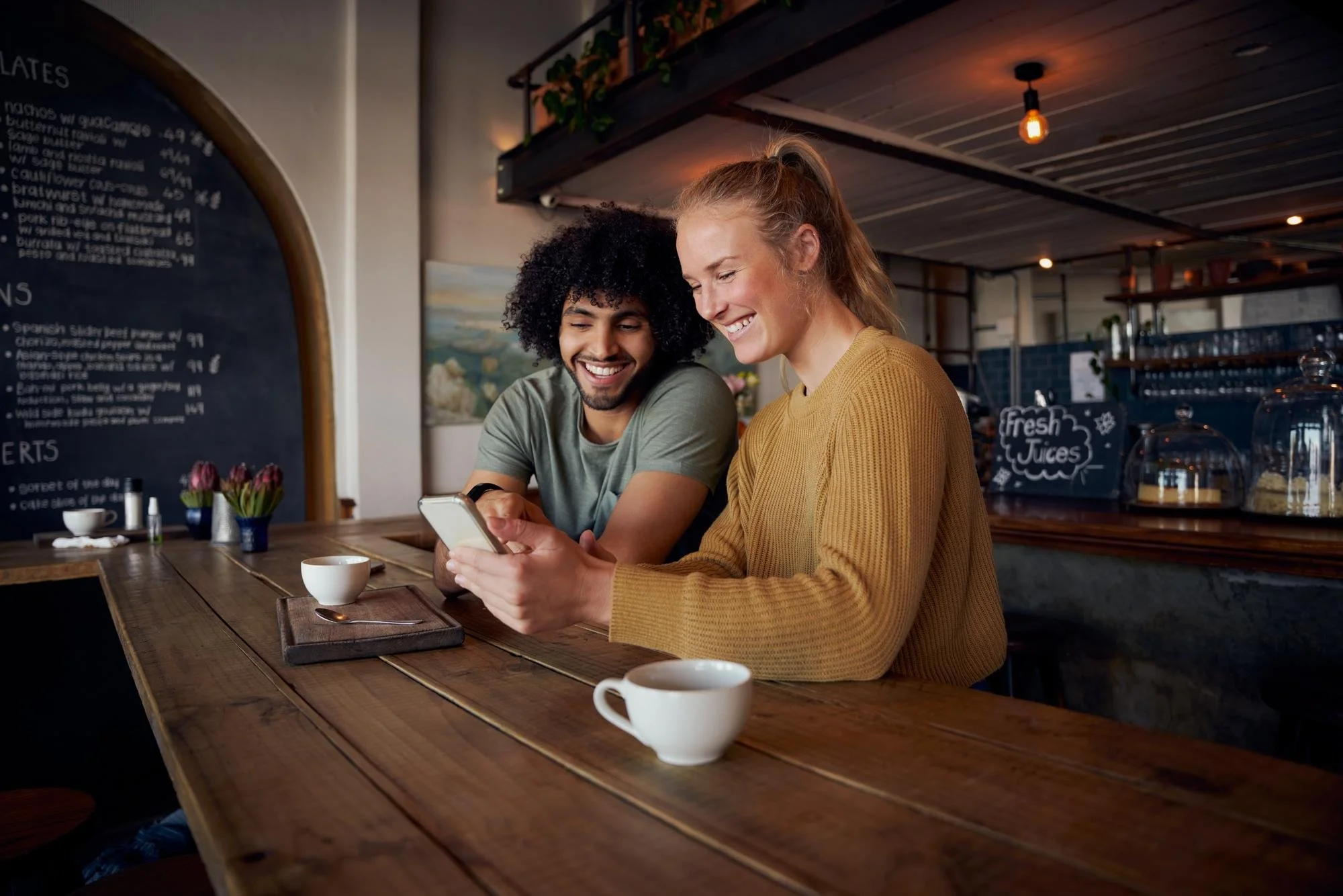 A man and a woman sitting at a wooden counter in a cafe, looking at a phone and smiling. There are cups on the counter, and a menu board and some flowers visible in the background. WINCHR HR services & consultation.