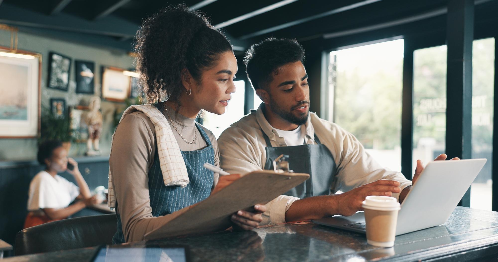 Two young people working together at a cafe, looking at a laptop. One is holding a clipboard, and the booking a HR Consultant in hospitality.