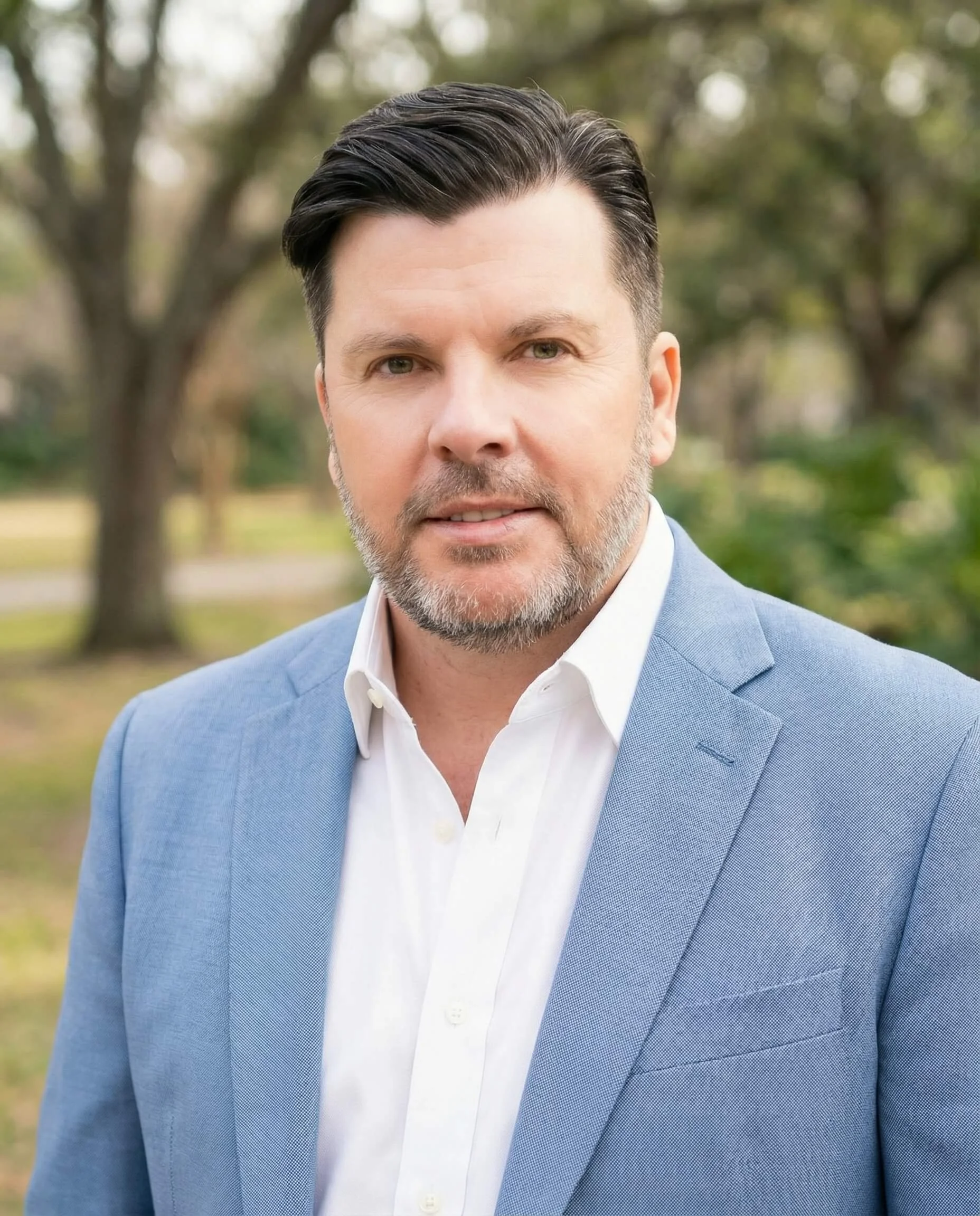 A man with dark hair and a beard, wearing a blue blazer and white shirt, standing outdoors with trees in the background.