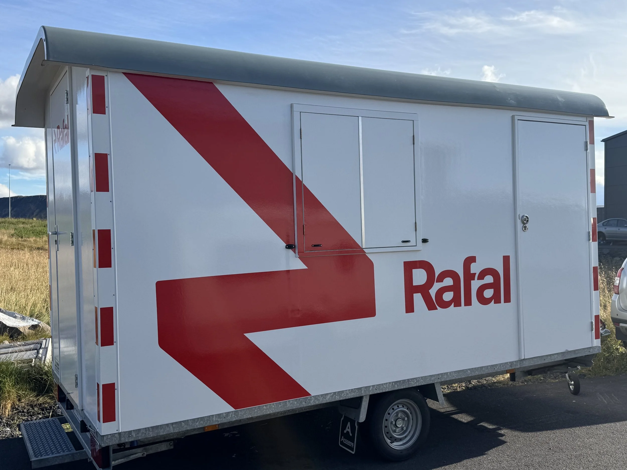 Hvítur vinnuskúr á hjólum með stóru rauðu myndmerki og heitinu Rafal á hliðinni.