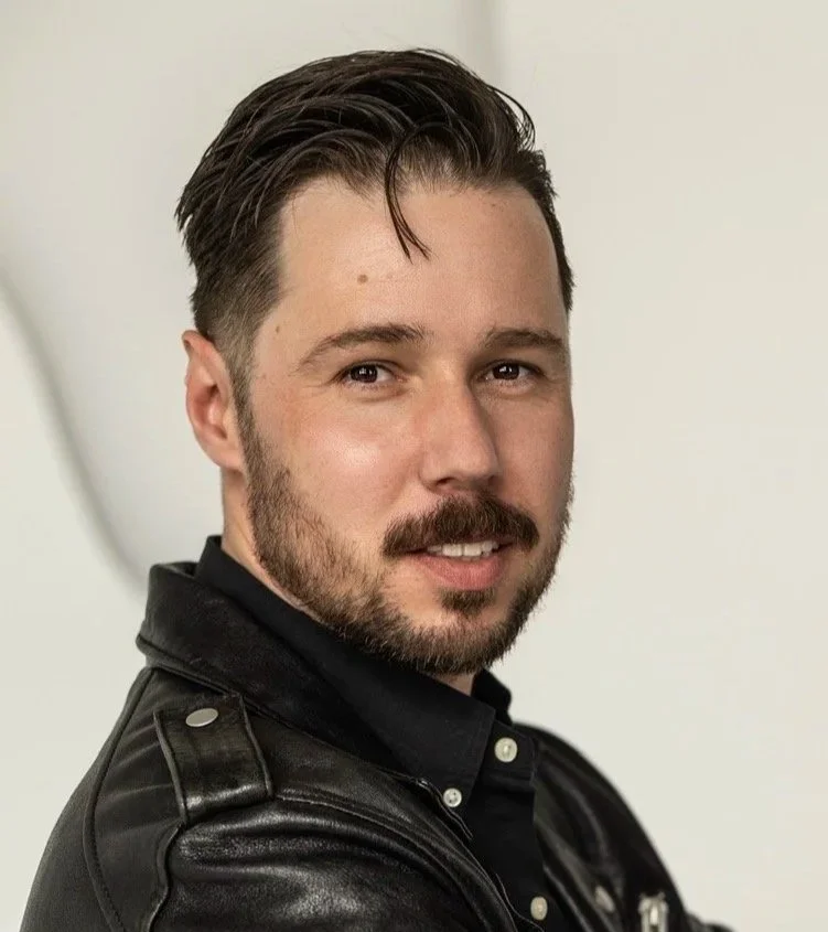 A young man with dark hair styled back, a mustache and beard, wearing a black leather jacket, smiling slightly.