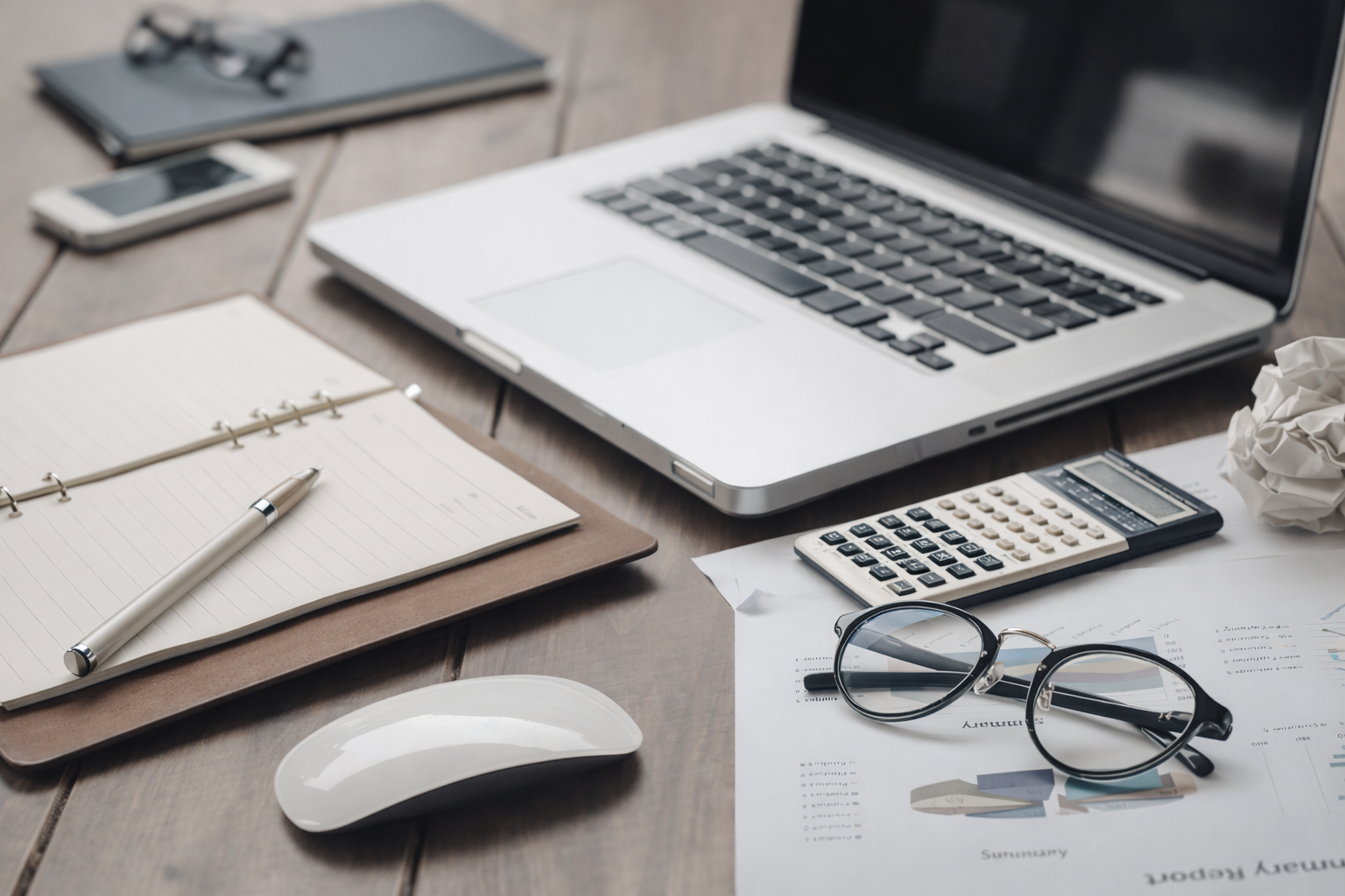 Arbeitsplatz mit Laptop, Notizbuch, Kugelschreiber, Smartphone, Taschenrechner, Brille, Papier und eine Schreibtischlampe.