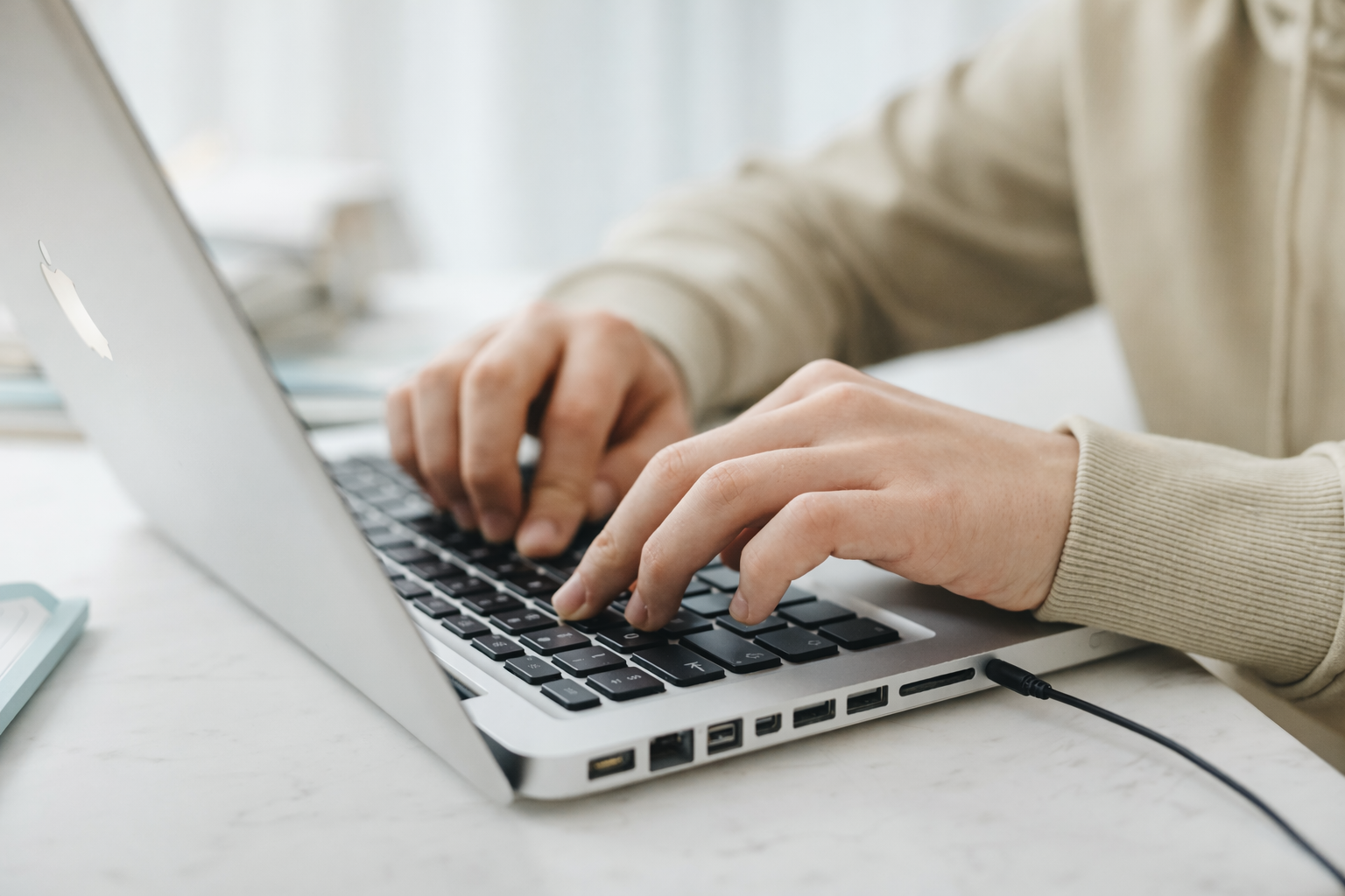 Nahaufnahme von einer Person, die auf einer silbernen Laptop-Tastatur tippt, auf einem weißen Schreibtisch, Kabel ist eingesteckt.