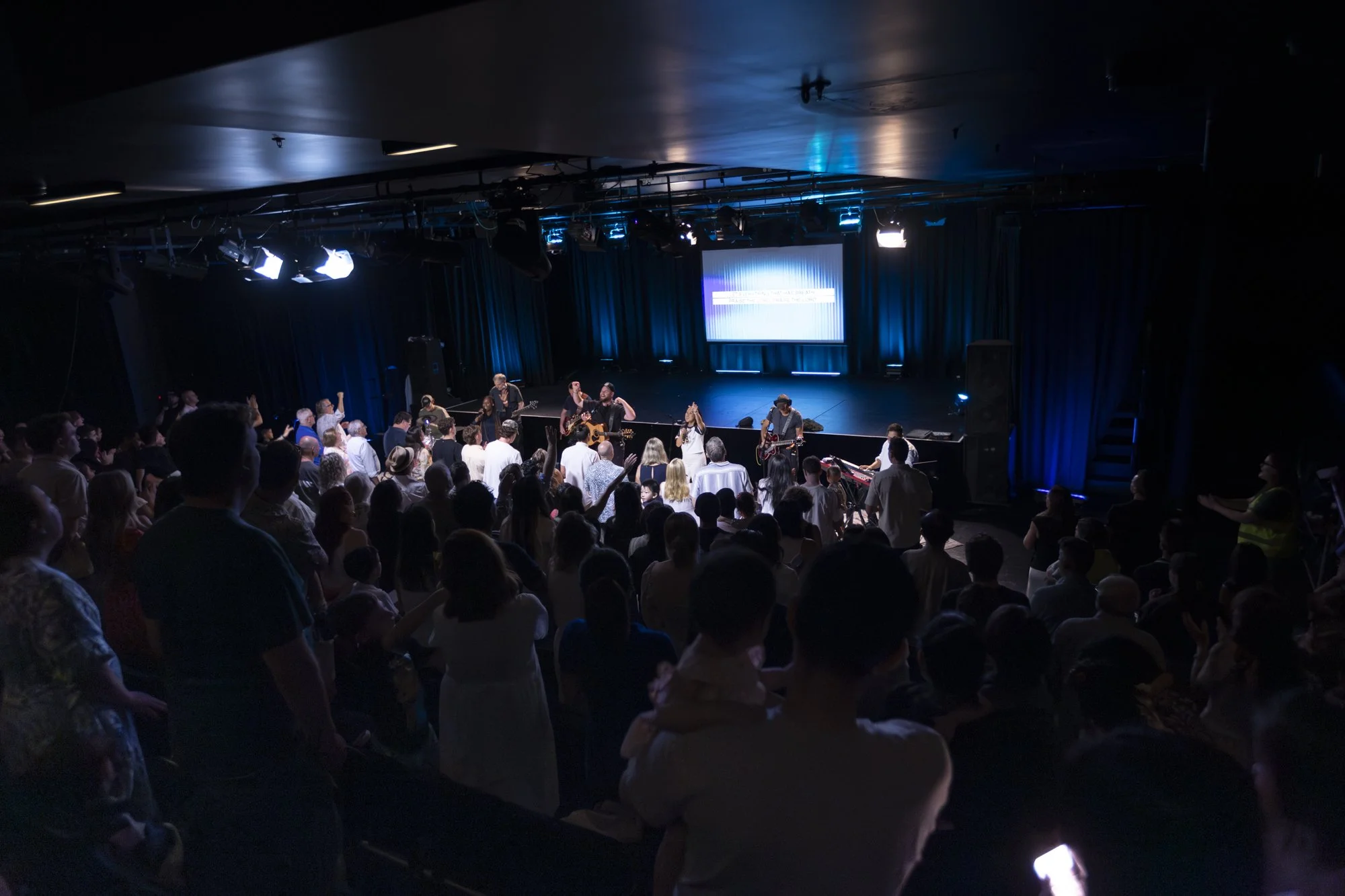 Audience standing and watching a live band performing on stage in a dimly lit concert venue.