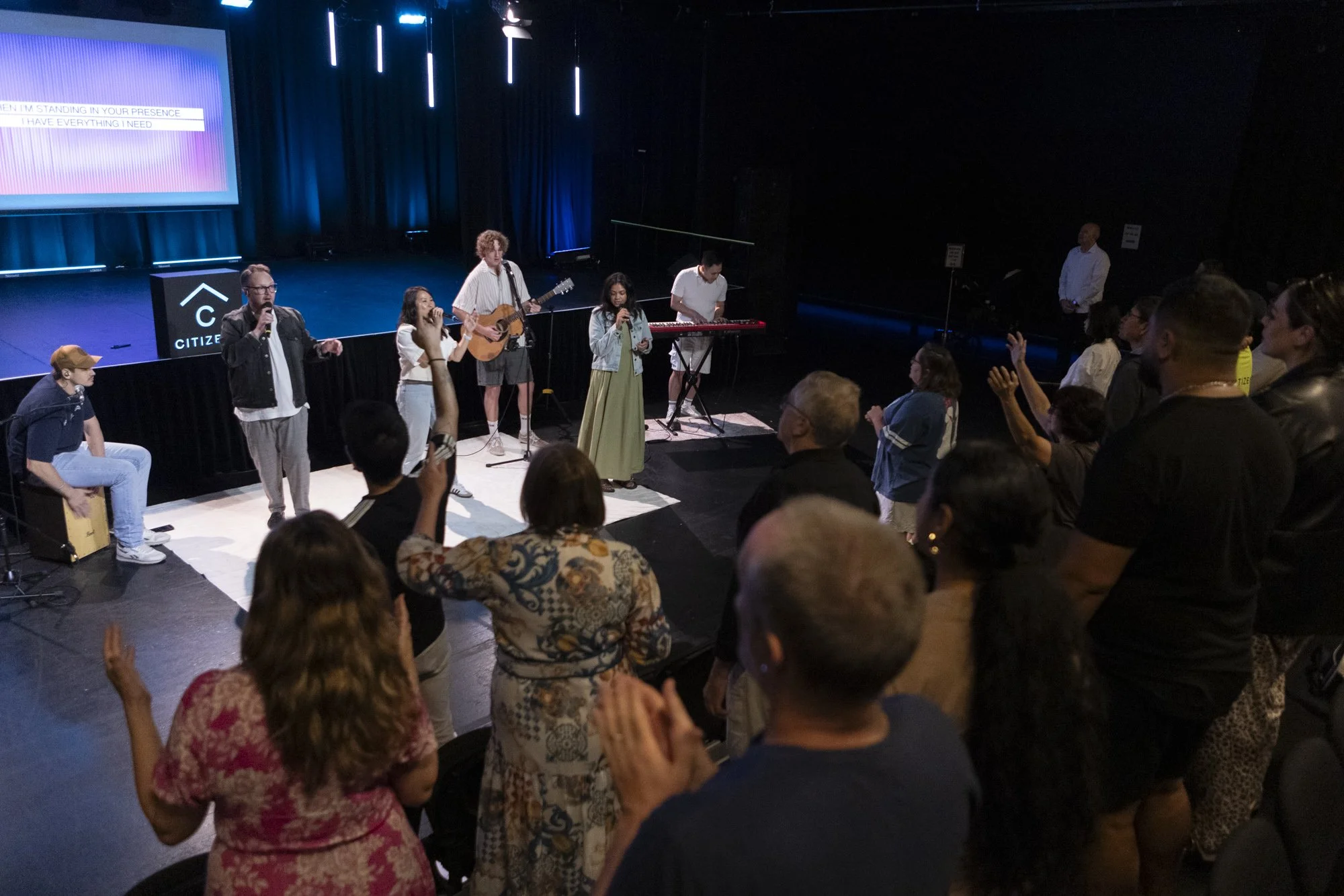 People standing and singing in front of an audience on a small stage in a dark room with blue lighting. Musical instruments include a guitar, a keyboard, and a cajón. Some audience members are clapping and raising their hands.
