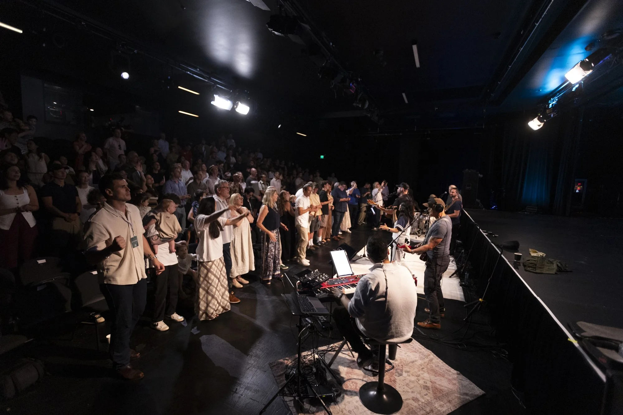 A group of people standing and participating in a religious or spiritual ceremony on a stage with musicians and microphones, in front of an audience in a dimly lit auditorium.