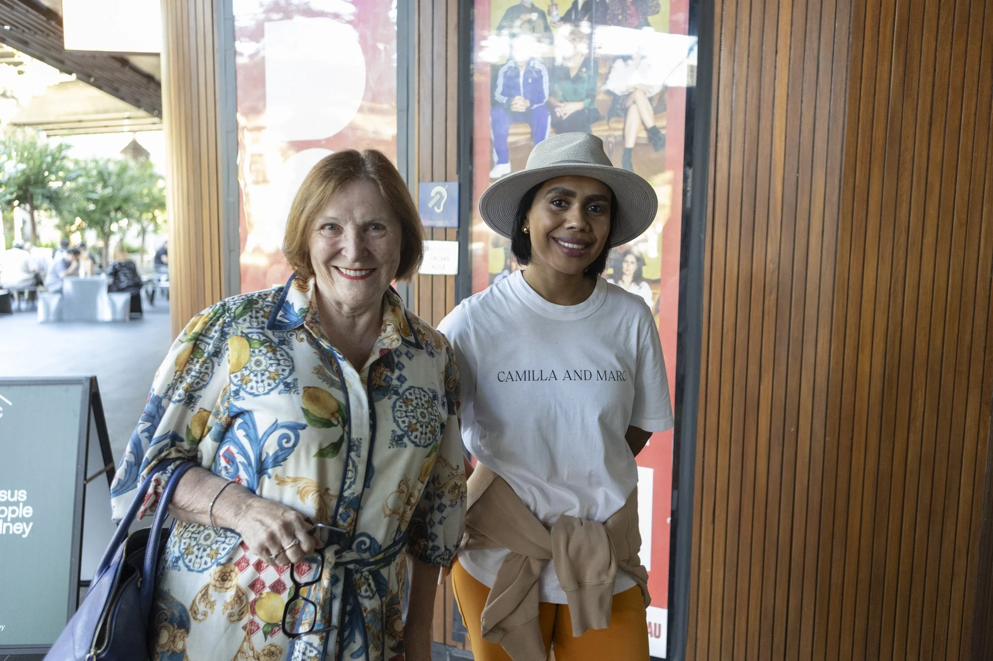 Two women smiling and standing indoors in front of a wooden wall. The older woman on the left has short, light brown hair and is wearing a colorful, patterned blouse and carrying a blue bag. The younger woman on the right has short, dark hair, a beige hat, a white T-shirt with the text "CAMILLA AND MARC," and mustard-colored pants.