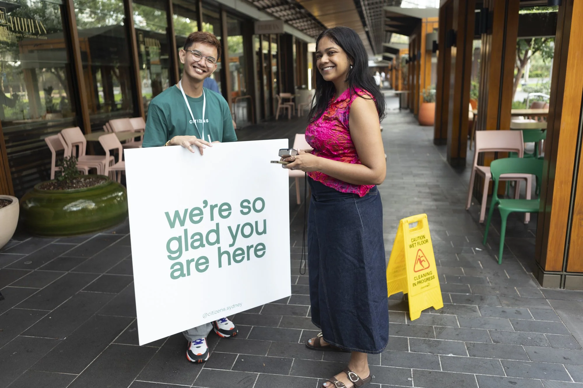 A young man and woman are standing outside a restaurant, smiling and holding a large white sign that reads 'we're so glad you are here.' The young man is wearing a green T-shirt and glasses, and the woman is dressed in a pink top and a long denim skirt. There is a yellow caution wet floor sign on the ground nearby.