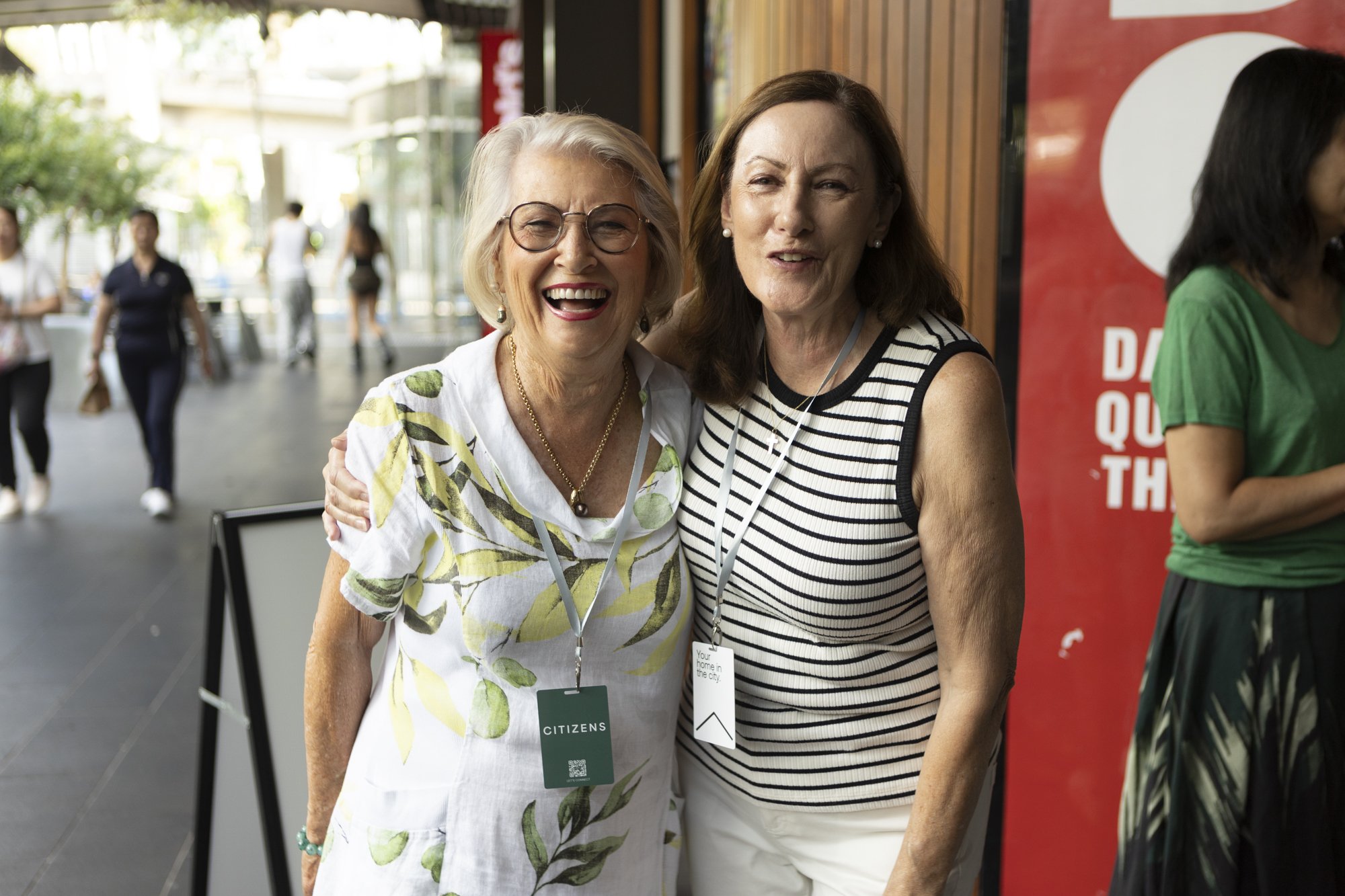 Two women smiling and posing for a photo outdoors at an event. One has blonde hair, glasses, and is wearing a white floral dress with a "Citizen" badge. The other has brown hair, is wearing a striped sleeveless top, and has a badge around her neck. They are standing close together with their arms around each other.