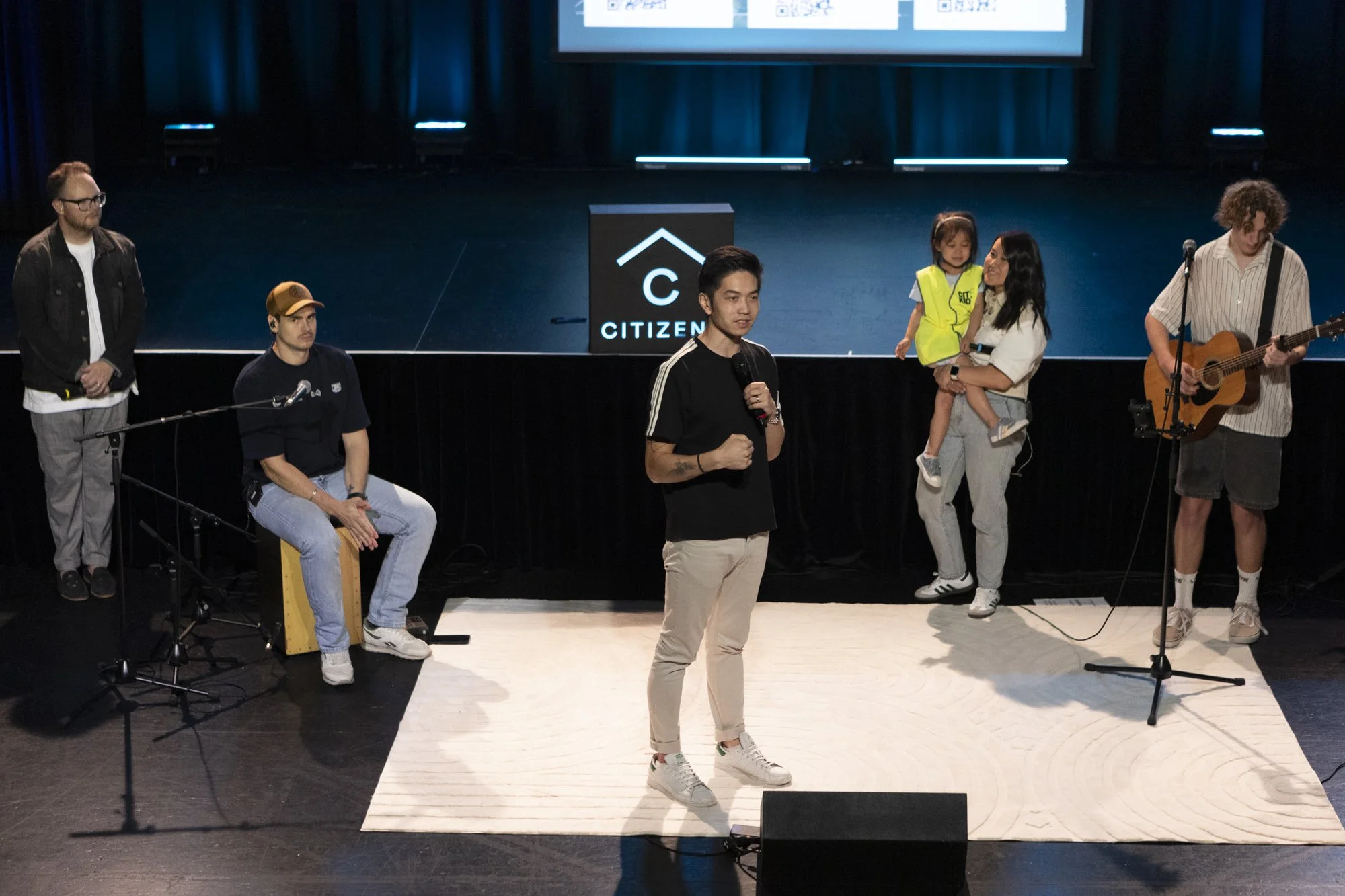 A group of five people on stage, with a man in the center speaking into a microphone. To his right, a woman is holding a young girl, both smiling. Two musicians are on the left and right sides, one playing a guitar and the other sitting on a cajón. There is a screen in the background with the logo of Citizen.