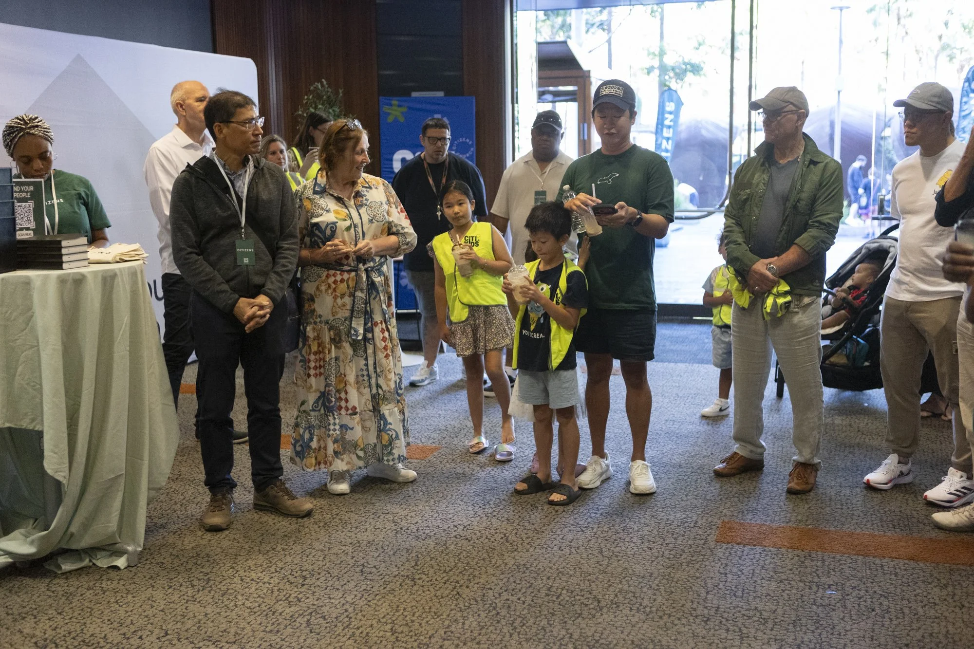 A group of people standing indoors, some holding drinks, with children in yellow safety vests, near a table with brochures, in a well-lit entrance area.