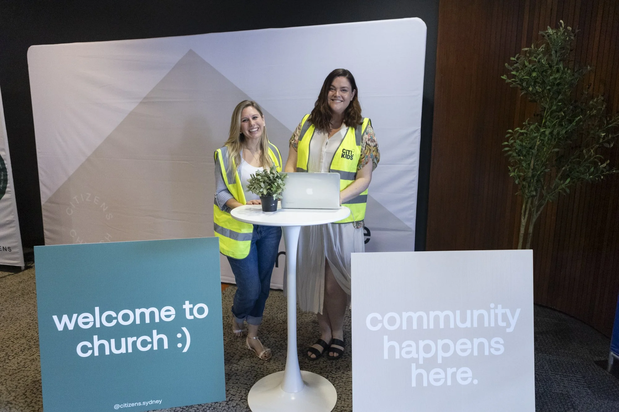 Two women standing behind a small high table with a laptop and a potted plant, at an event with signs that say "welcome to church :)," and "community happens here," wearing reflective safety vests, smiling, with a backdrop and a potted plant nearby.