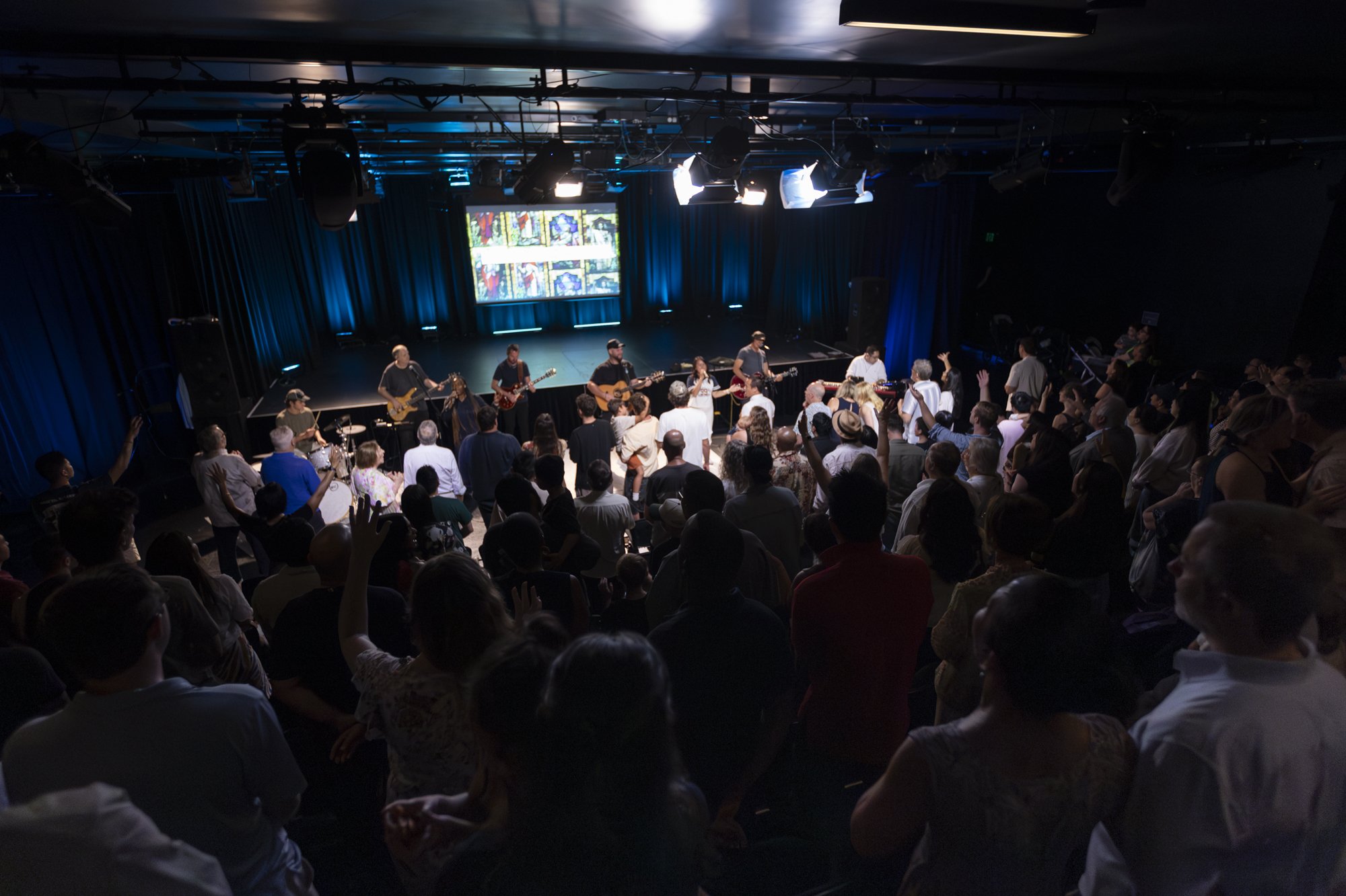 Concert performance on a stage with multiple musicians playing guitars and drums, illuminated by stage lights, with an audience watching in a dark room.