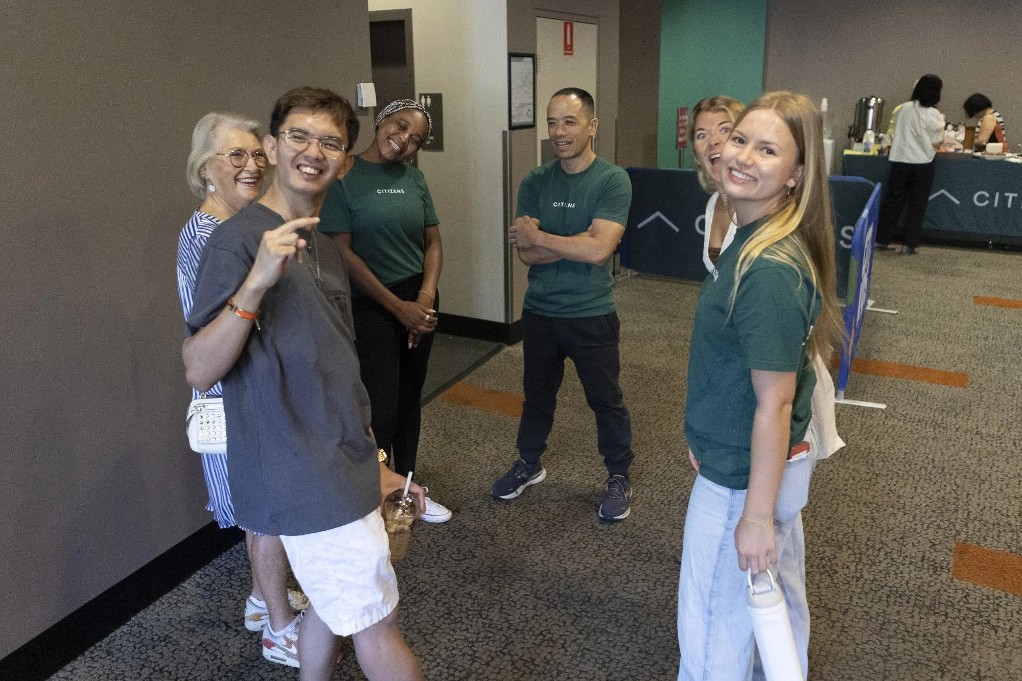 Six diverse people, smiling and engaging in conversation, standing in a hallway near a refreshment area in a conference or event setting.